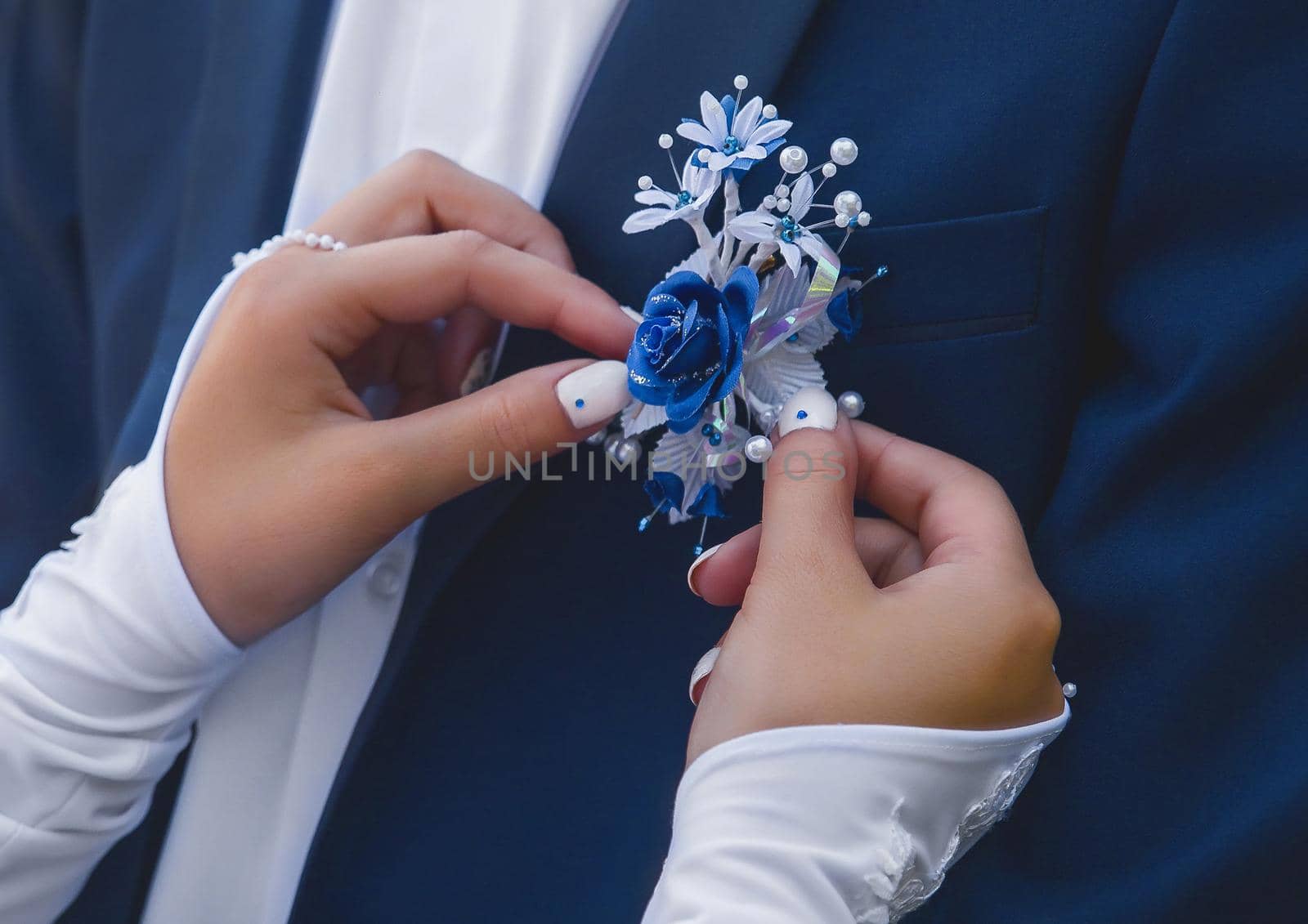 Hands of the bride in wedding gloves touch the decoration in the form of a rose flower on a blue suit of the groom, close-up.