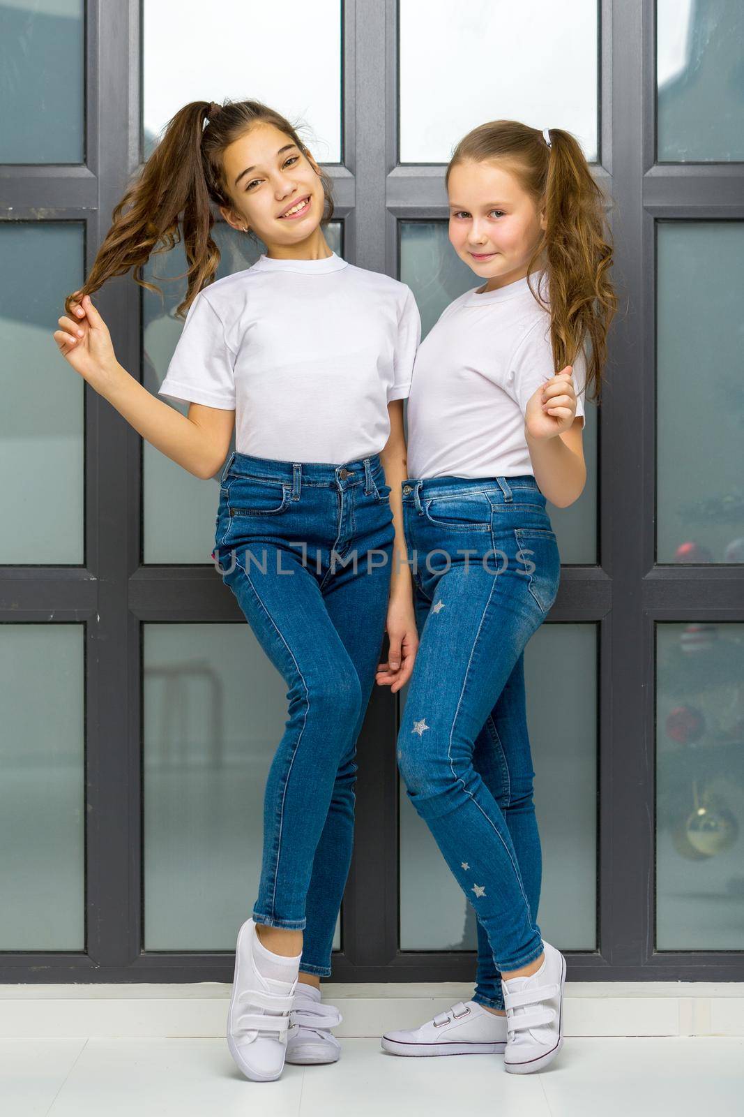 Two beautiful little girls of school age posing in the studio near a large panoramic window. Outside the window it is already evening, dark, the concept of tenderness and beauty.