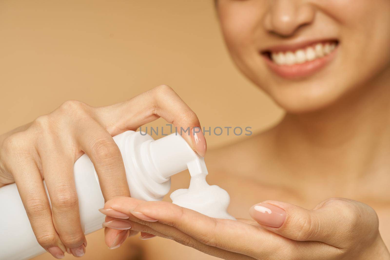 Cropped shot of young woman smiling, holding a bottle of gentle foam facial cleanser isolated over beige background. Beauty products and skin care concept