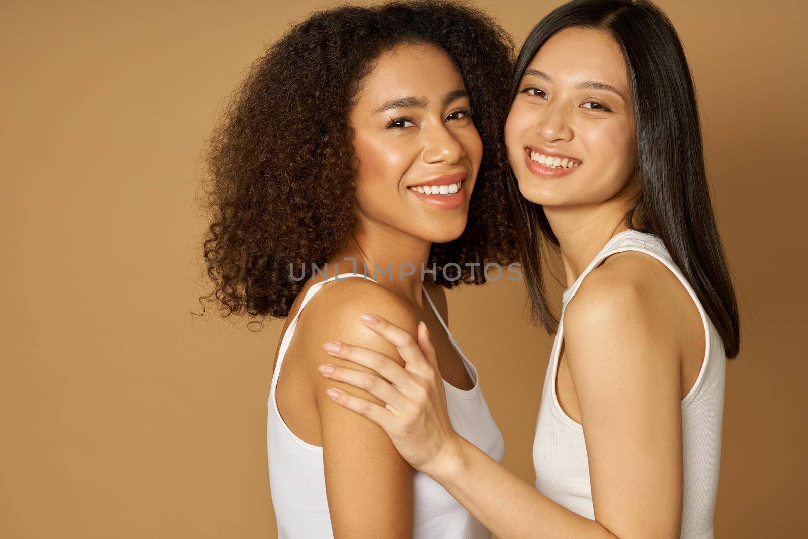 Portrait of two stunning mixed race young women with perfect skin smiling at camera while posing together, standing isolated over light brown background by friendsstock