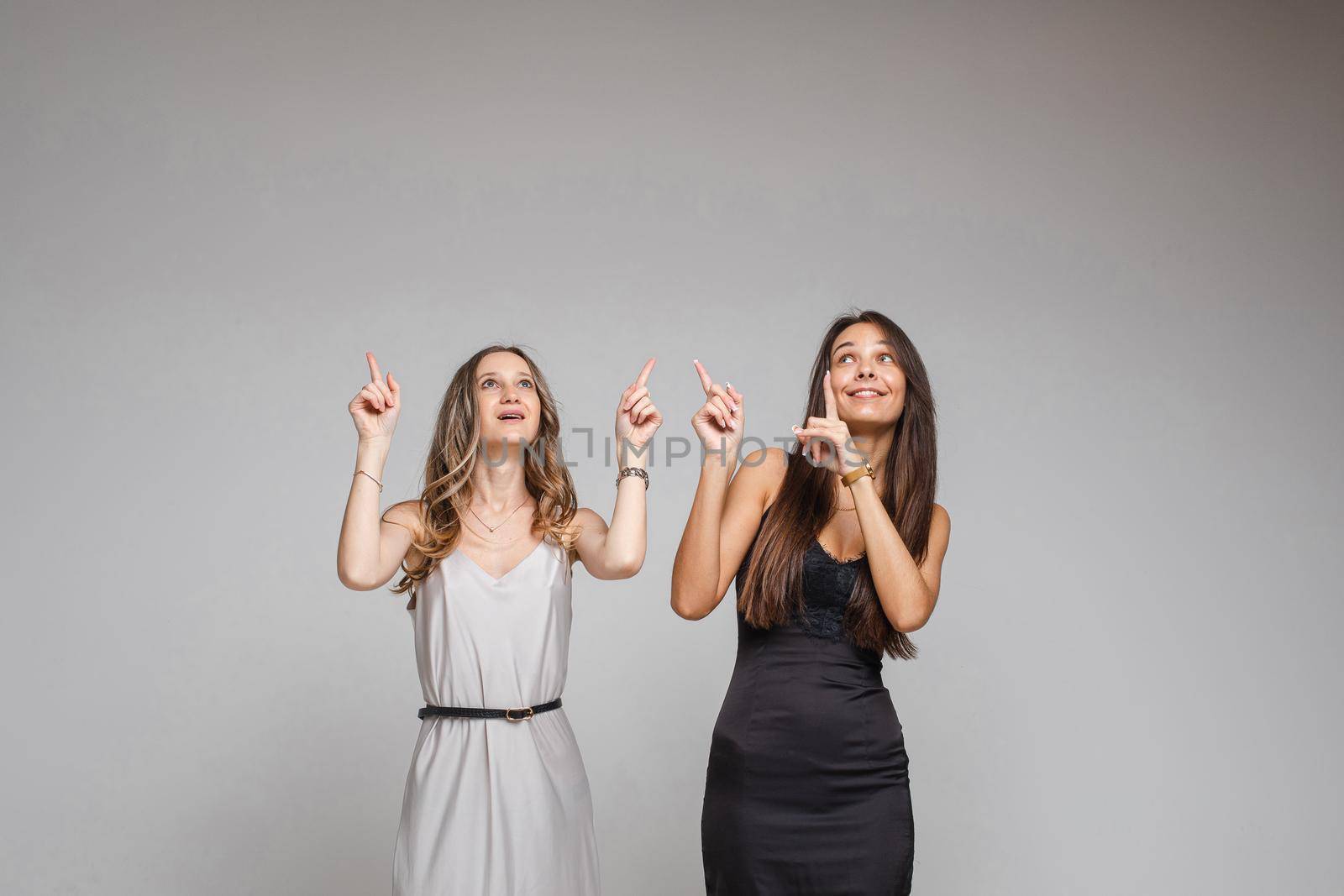 Two pretty women standing in studio pointing their fingers up, isolated on grey background. Party concept. Copy space