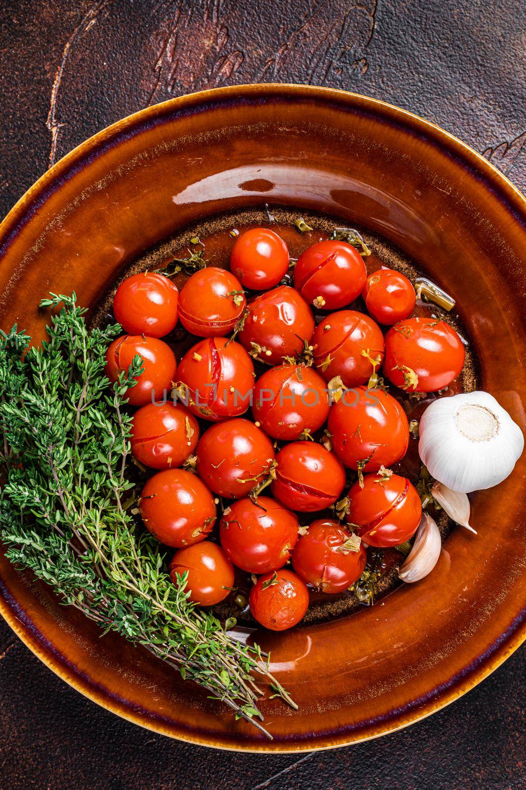 Pickled cherry tomatoes in a rustic plate with garlic and thyme. Dark background. Top view. Copy space by Composter