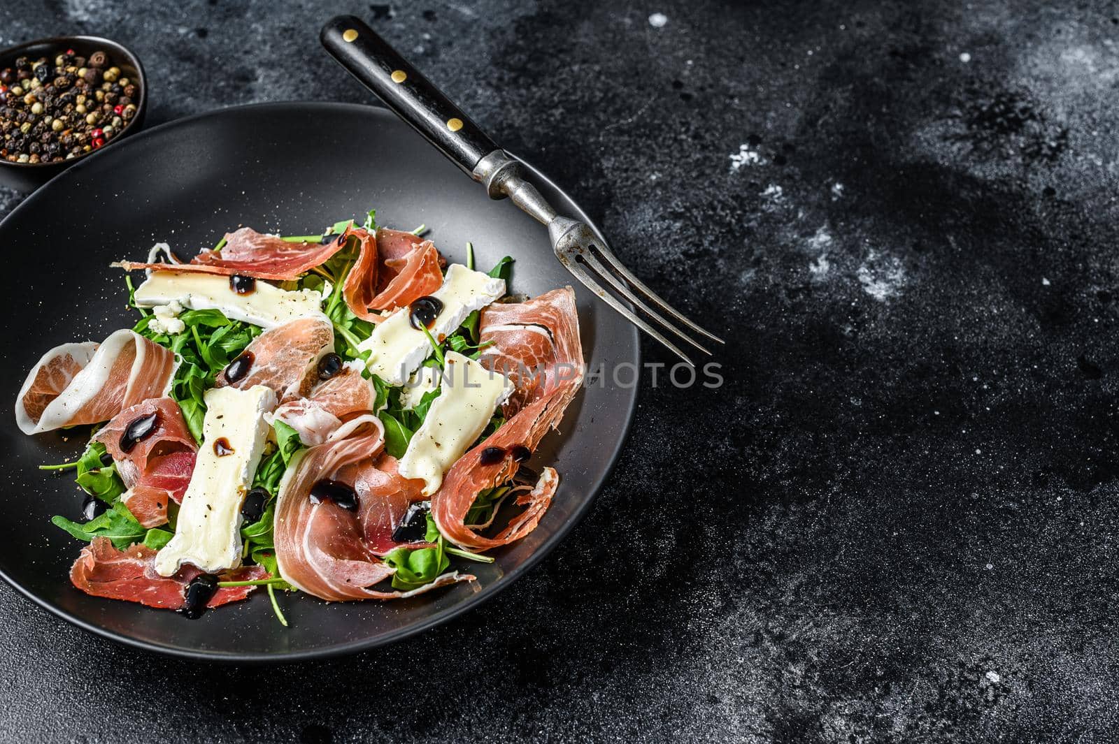 Prosciutto crudo ham salad with brie camembert cheese and arugula on a plate. Black background. Top view. Copy space.