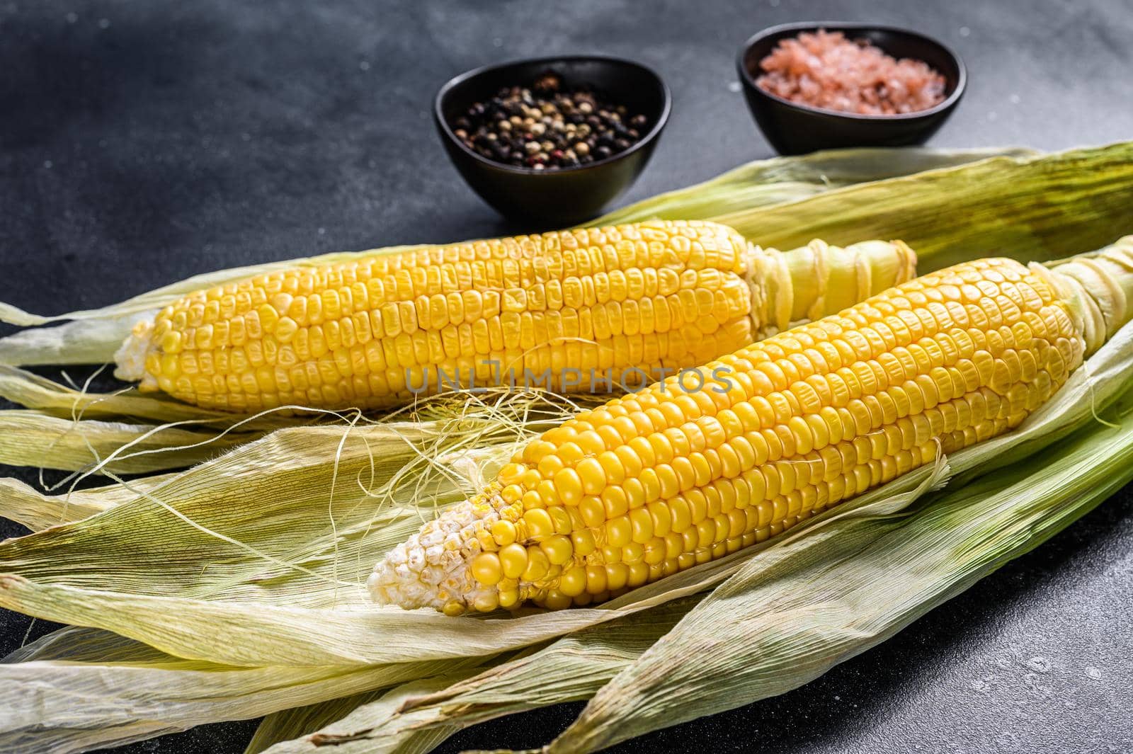 Cooked corn on cobs with salt and pepper. Black background. Top view.