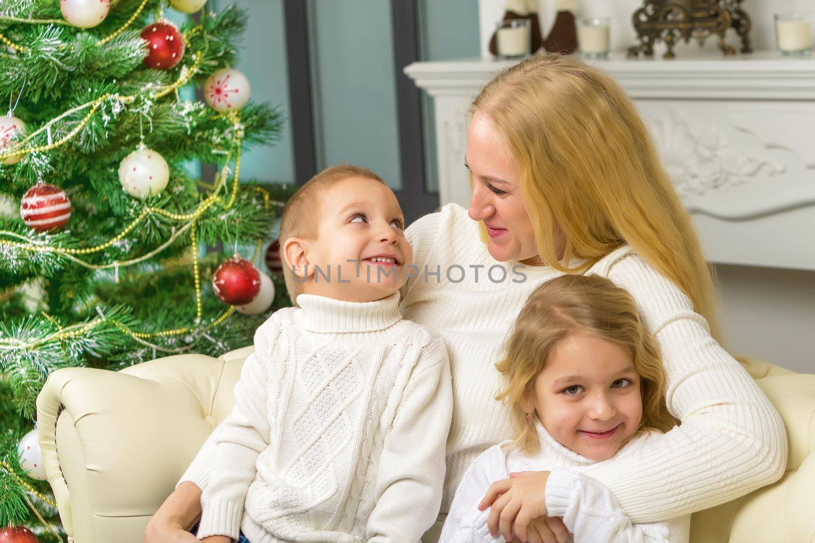 Happy Family Sitting on the Sofa in Christmas Interior with Decorated Fir Tree and Fireplace, Mom Hugging her Cute Little Kids, Her Son and Daughter Looking Lovingly at Her