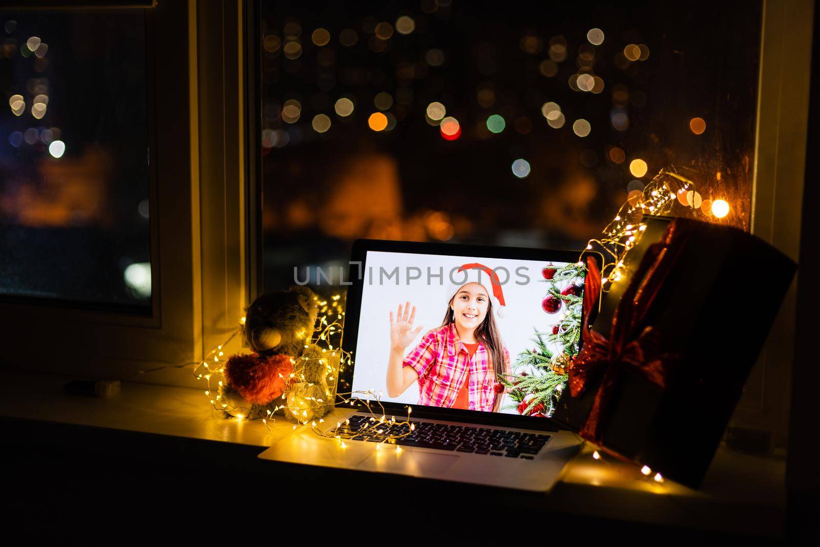 Laptop computer standing on table with gift and Santa hat in cozy room with little girl on the screen smiling, wishing Merry Christmas and Happy New Year by Andelov13