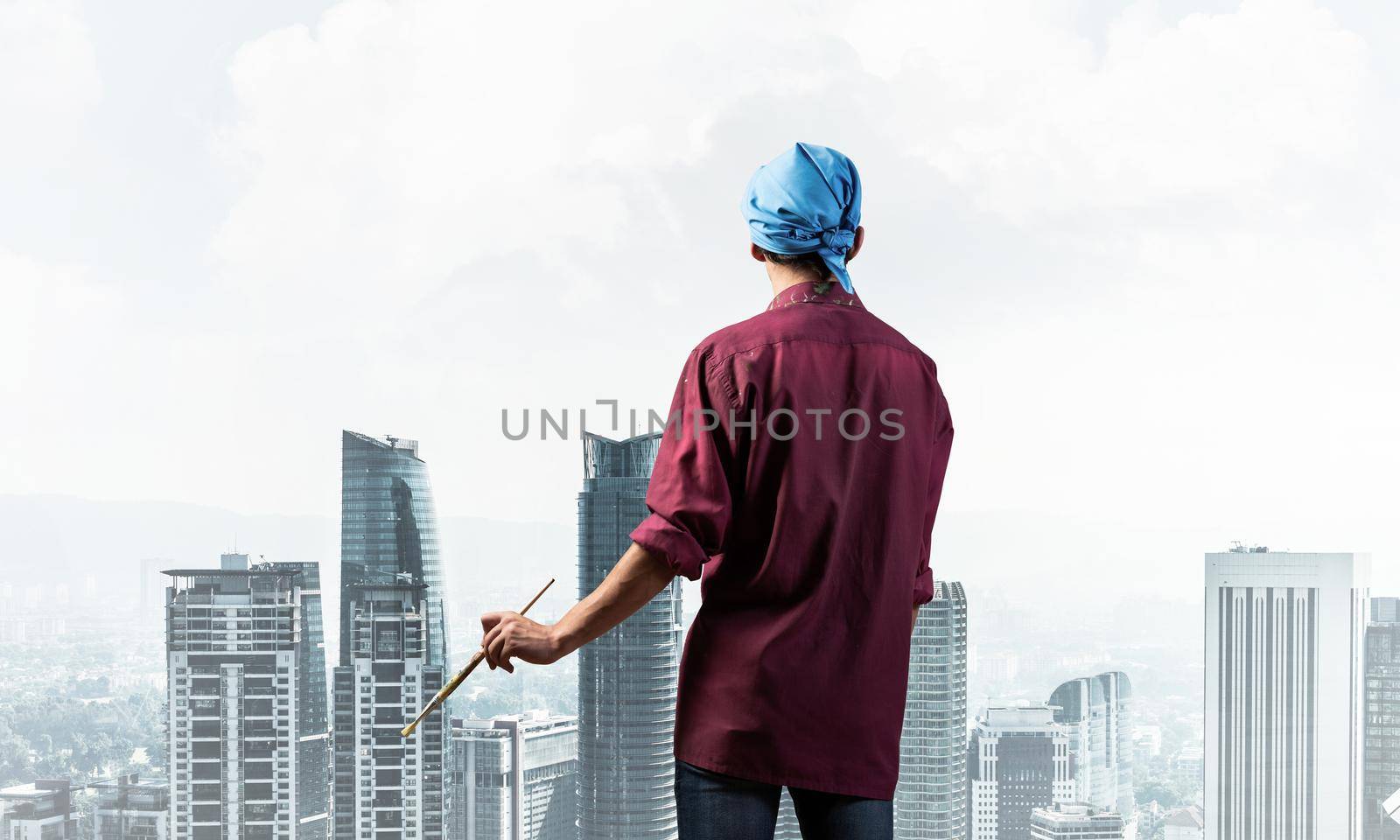 Young male painter artist holding paintbrush. Back view of painter in shirt and bandana on background cloudy sky and cityscape. Creative hobby and artistic occupation. Art school courses concept.