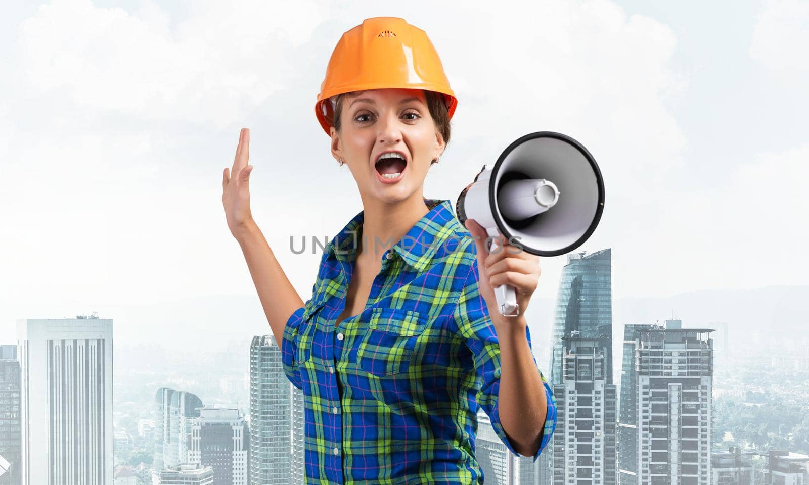 Expressive woman in safety helmet shouting into megaphone. Portrait of young emotional construction worker with wide open mouth on cityscape background. News announcement and advertisement.