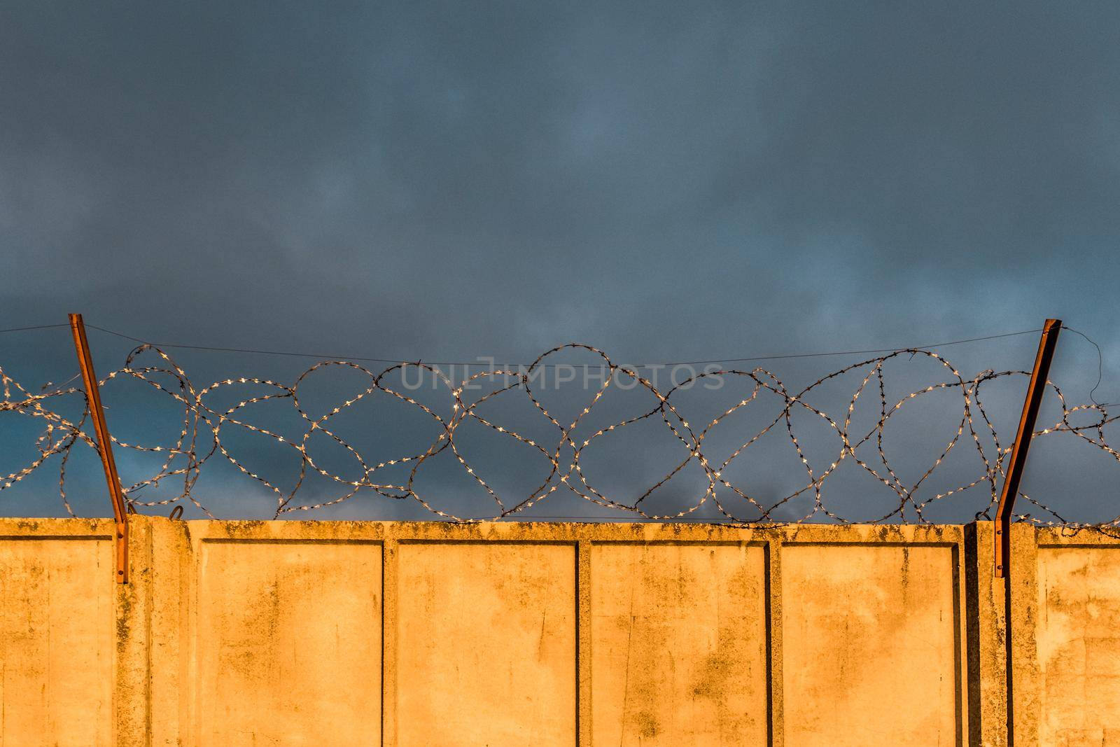 Barbed metal wire fence with concrete fence of a prison or closed object against a cloudy blue sky and sunset.