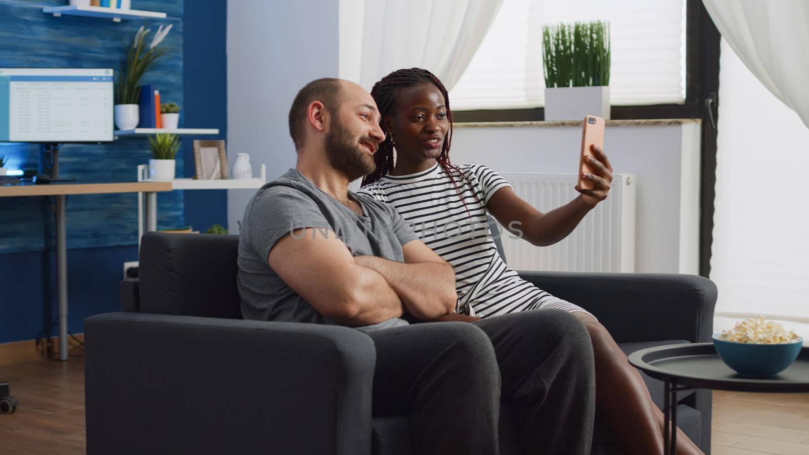 Interracial partners using video call on smartphone in living room. Cheerful multi ethnic couple chatting on online conference via internet for remote communication at home on couch