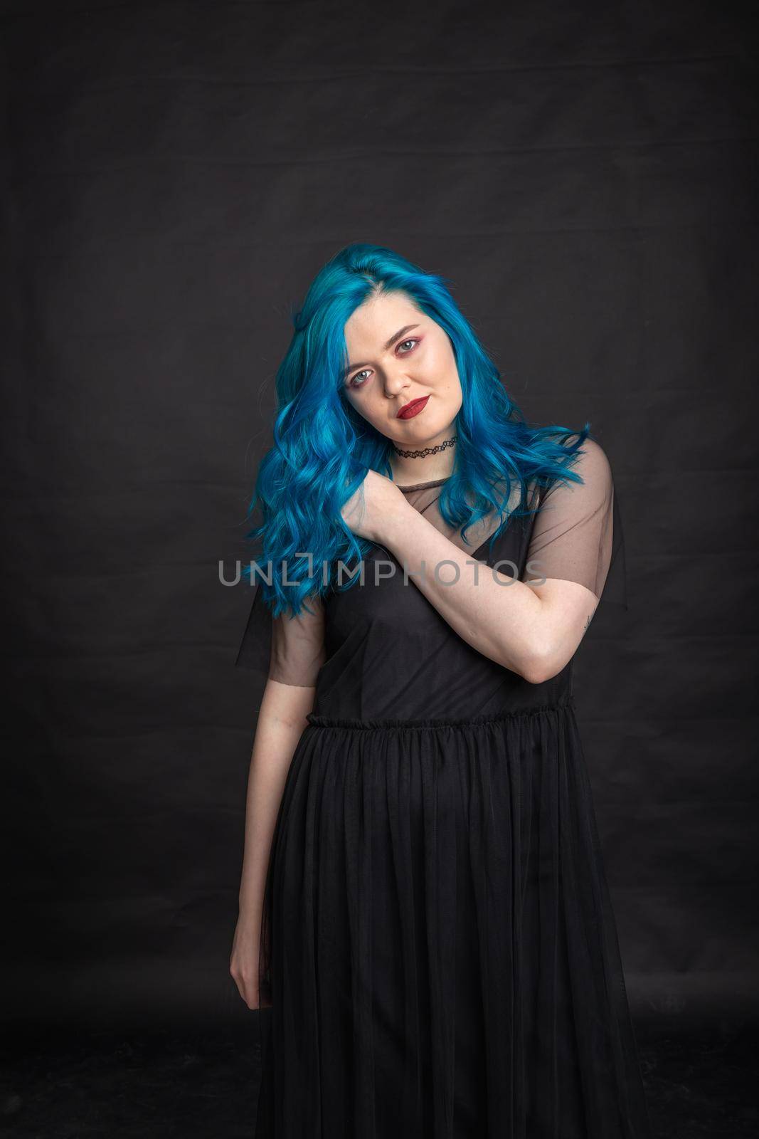 People and fashion concept - Woman with blue long hair dressed in black dress posing over black background.