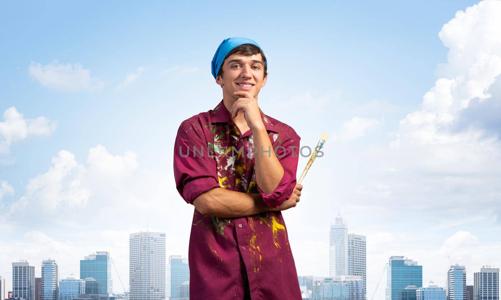Smiling young painter artist holding paintbrush. Portrait of happy art school studentin shirt and bandana on cityscape background. Artistic occupation and creative profession. Art courses promotion