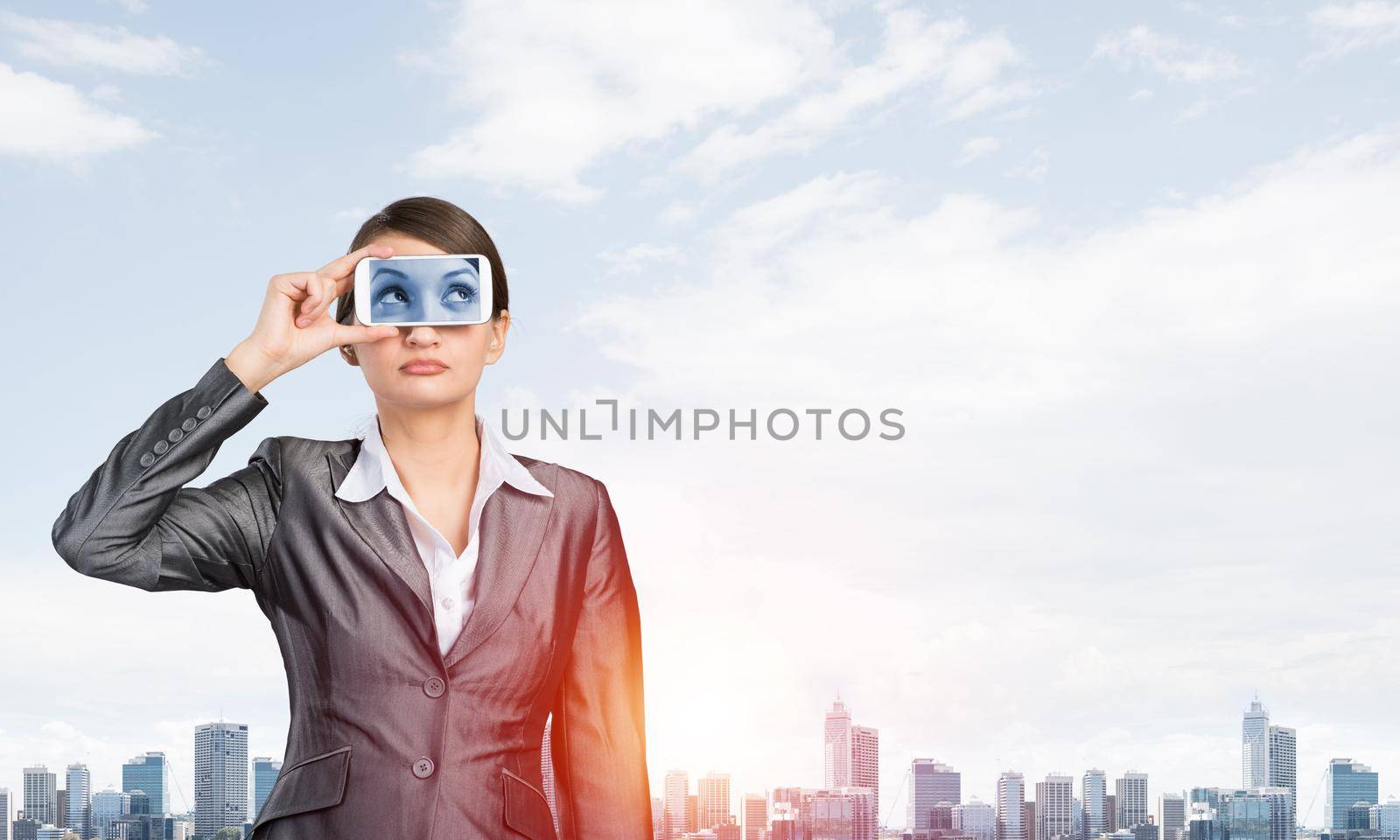 Portrait of pretty woman covering her eyes with smartphone. Businesswoman showing mobile phone with eyes on screen. Corporate businessperson on cityscape background. Mobile communication layout.