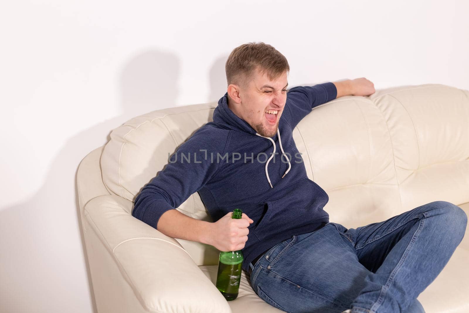 Depression and alcoholism concept - aggressive young man sitting on a sofa with bottle of alcohol.