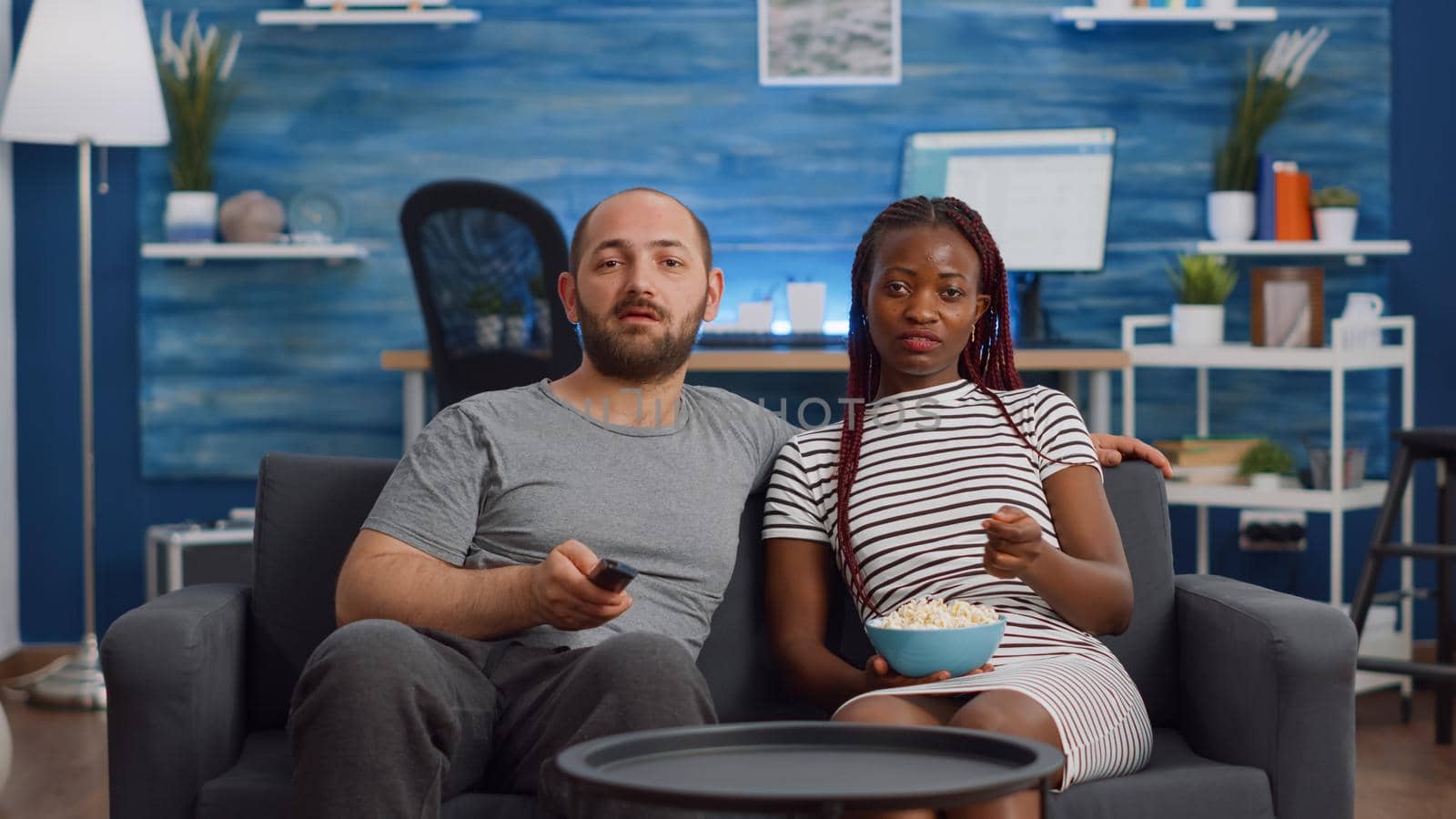 Young interracial couple watching television in living room. African american woman eating popcorn while caucasian man switching channels with TV remote control. Mixed race relationship