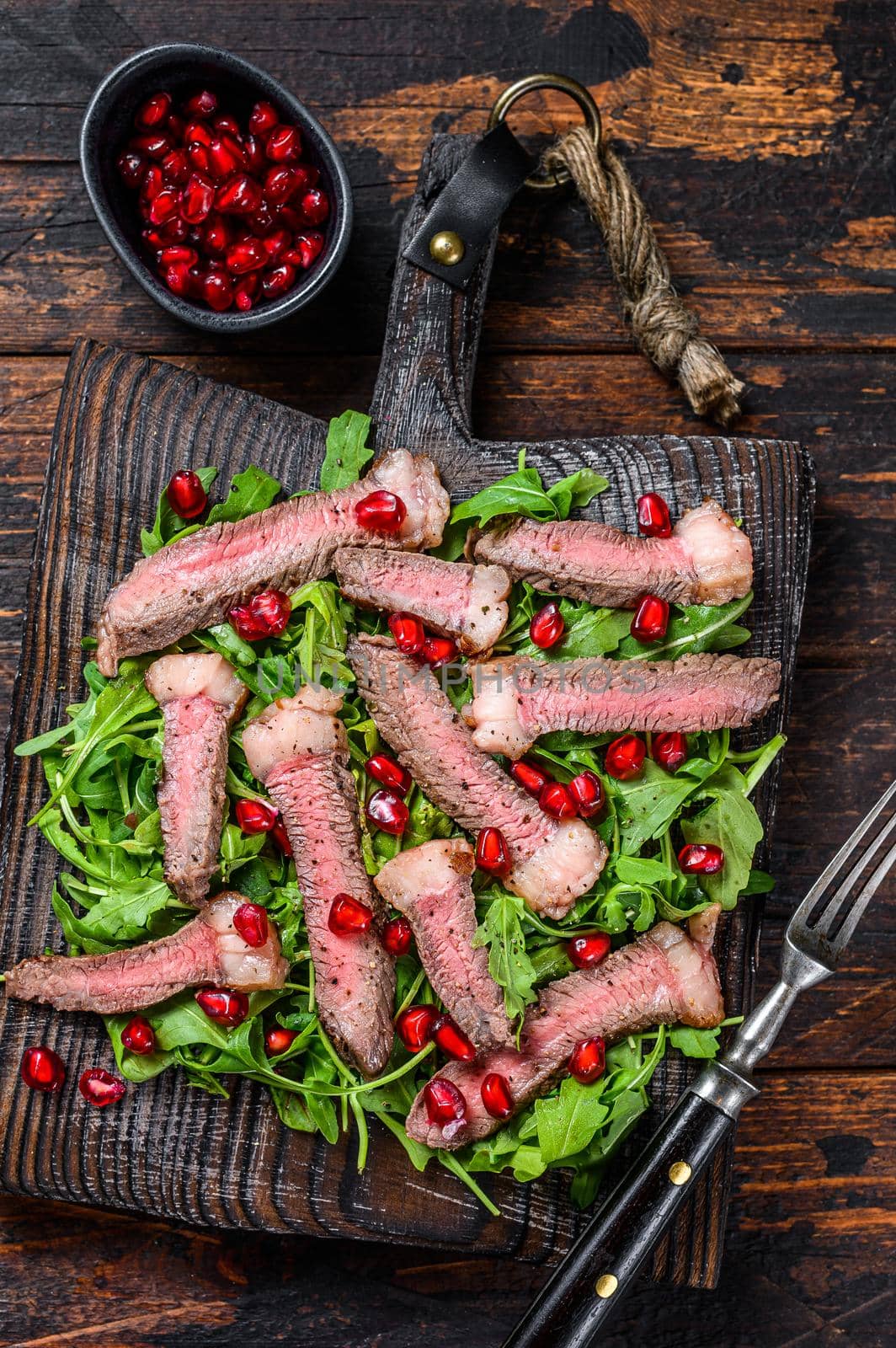 Grilled Beef Steak salad with arugula, pomegranate and greens vegetables. Dark wooden background. Top view by Composter