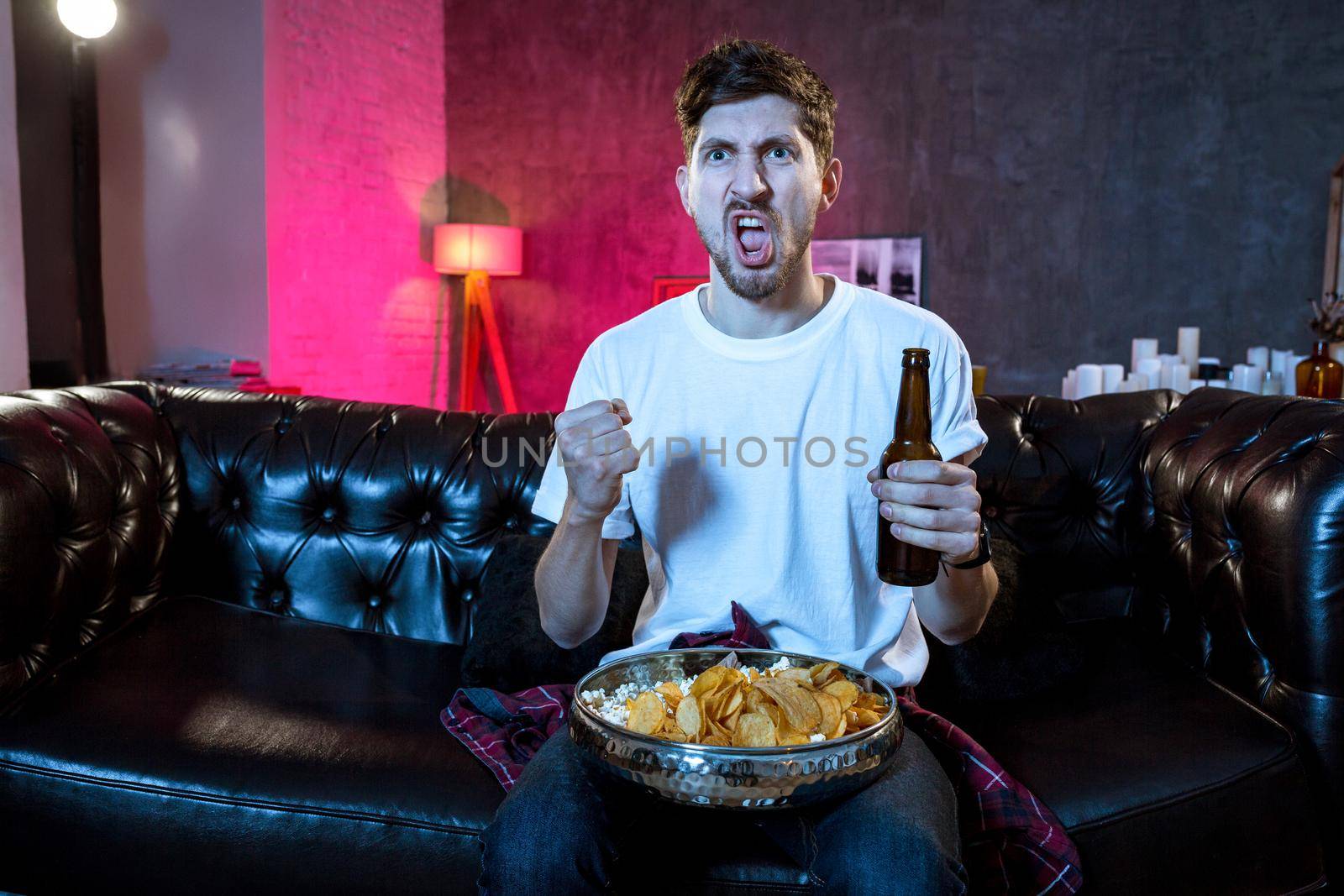 Young supporter man watching football game on television sitting at home couch with ball beer and chips in stress dejected and disappointed for failure shouting and complaining. Emotional fan watching the game of your favorite team