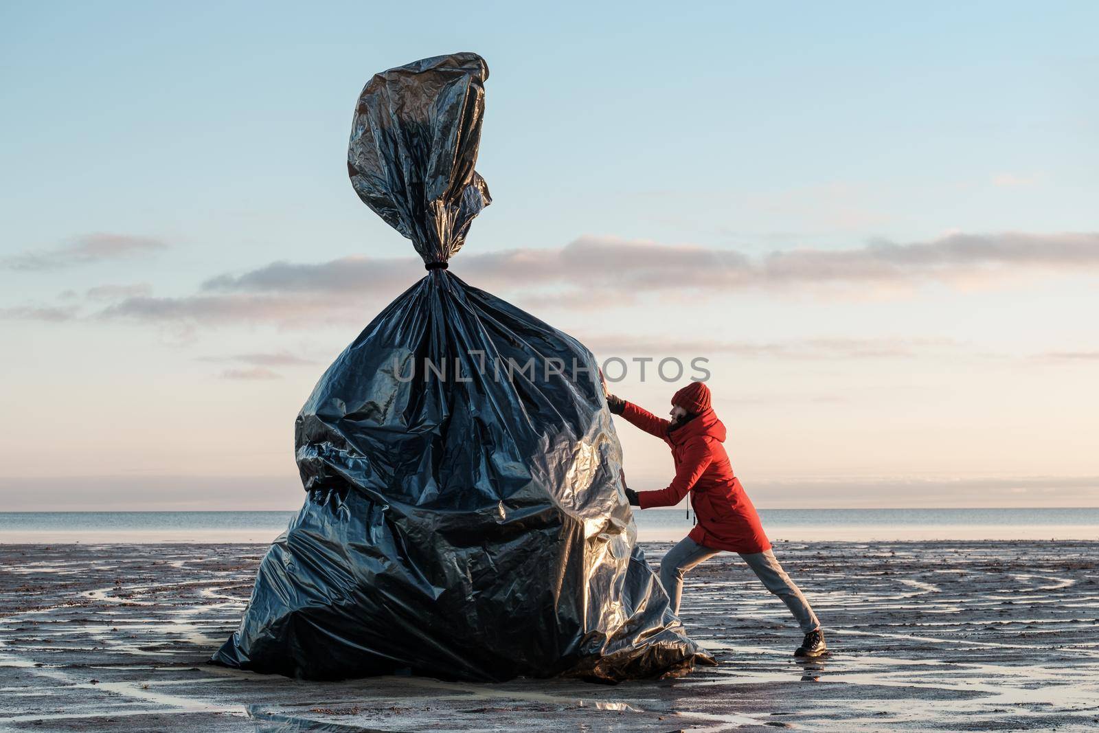 A woman pushes a huge black trash bag on the shore by vollirikan