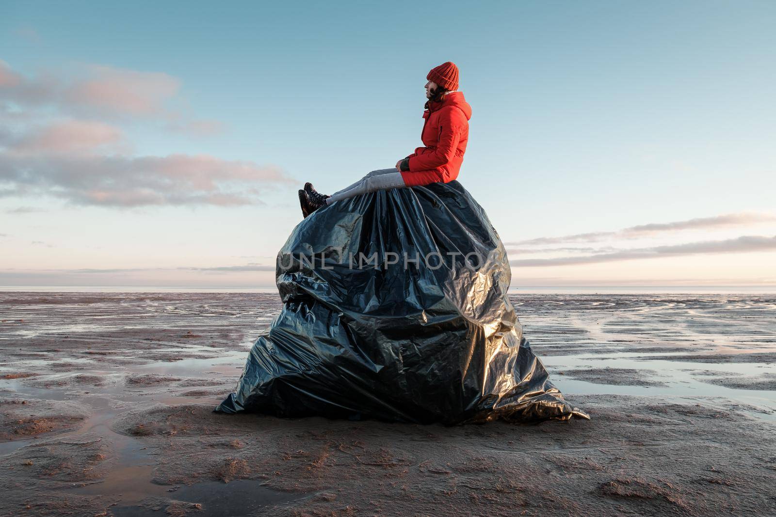 A woman sits on a huge black garbage bag on the shore. Environmental pollution by plastic. Recyclable packaging.