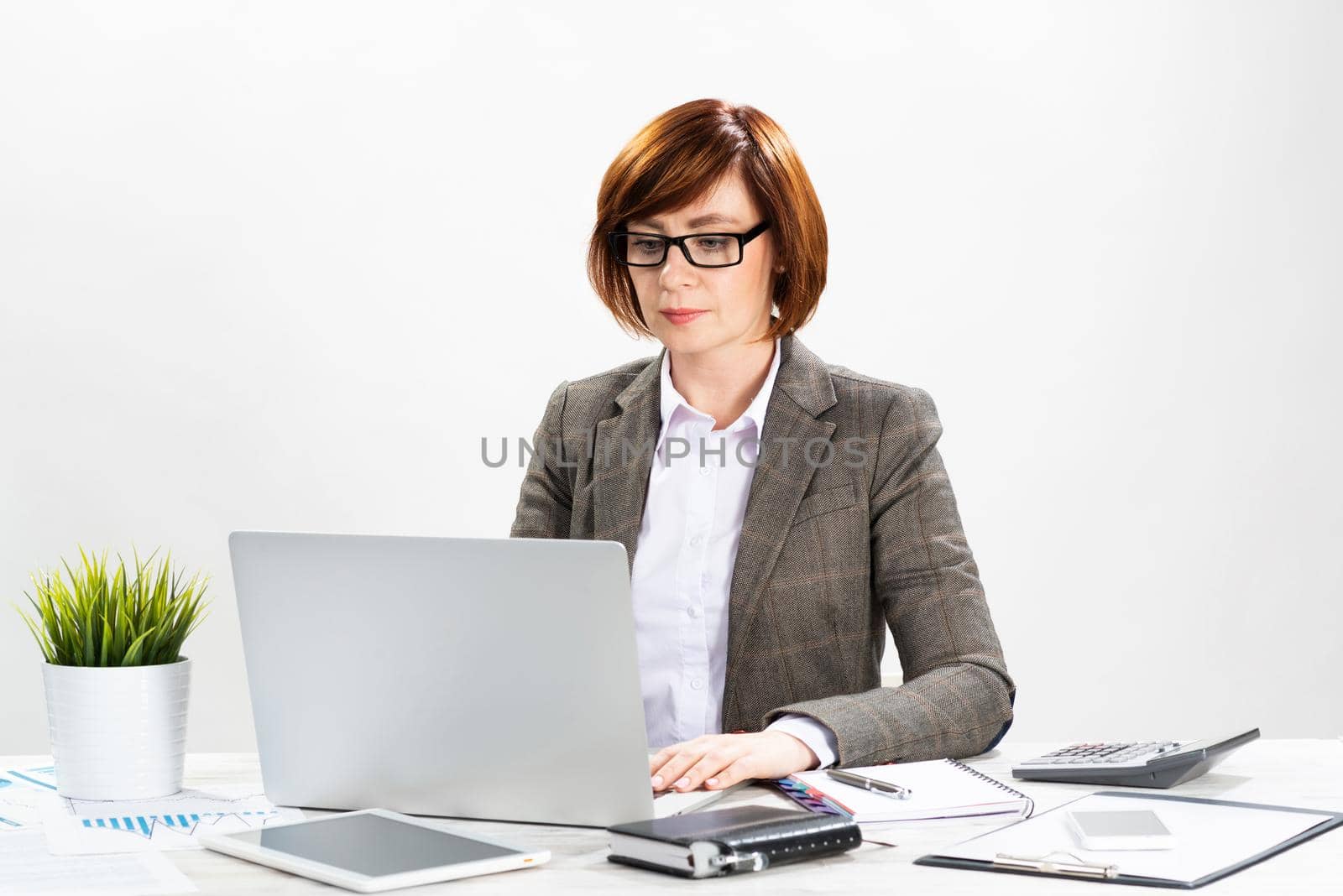 Attractive middle aged businesswoman working with computer in office. Business lady sitting at desk on white background. Successful corporate woman in suit and glasses posing at her workplace.