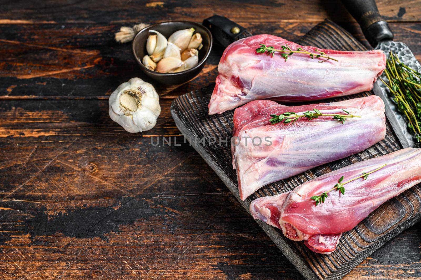 Raw lamb shanks meat on a cutting board with herbs. Dark wooden background. Top view. Copy space.