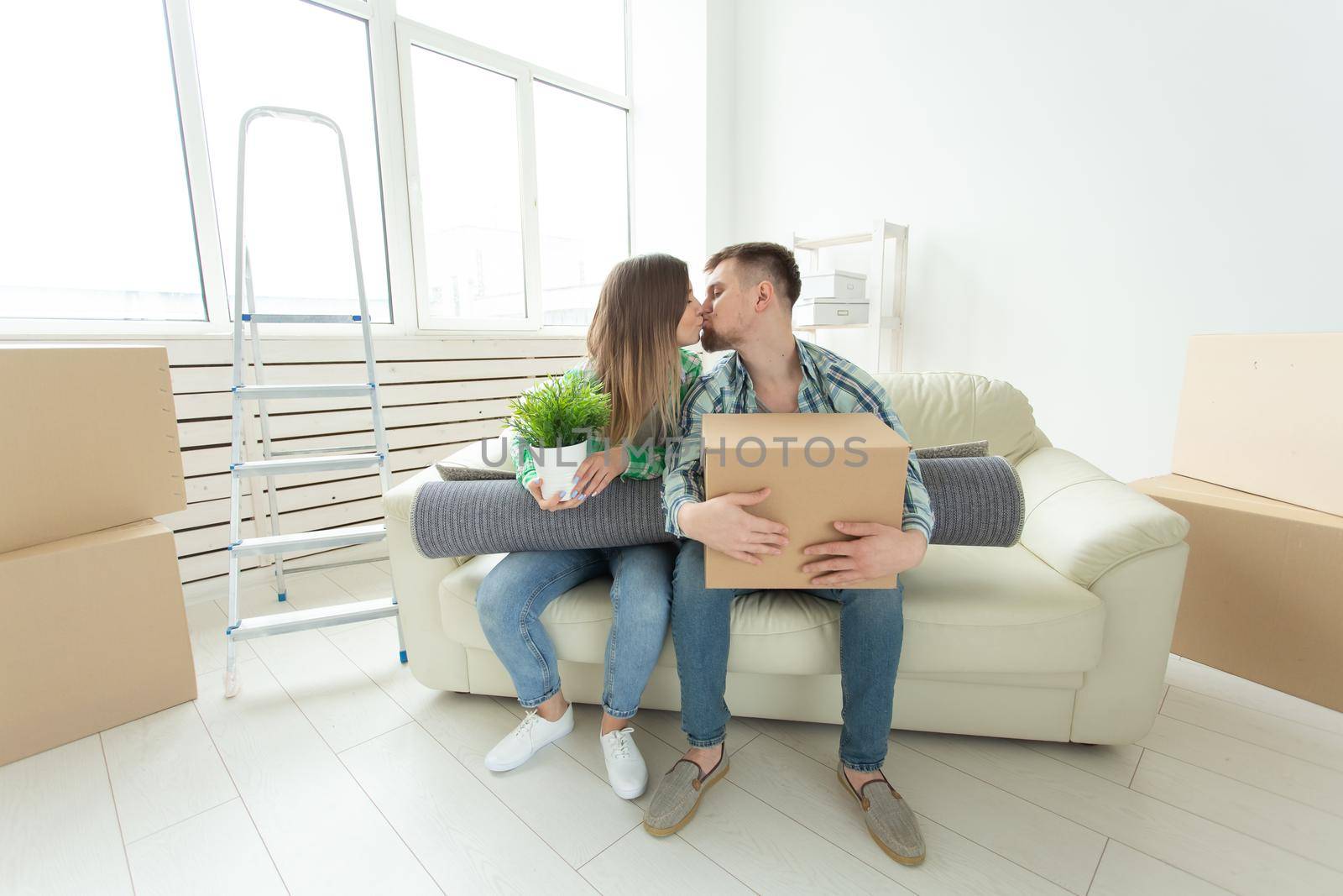 Charming young couple in casual clothes holding things in hands and sitting on sofa kissing to congratulate each other with housewarming. Concept of mortgage and housing affordability for young people