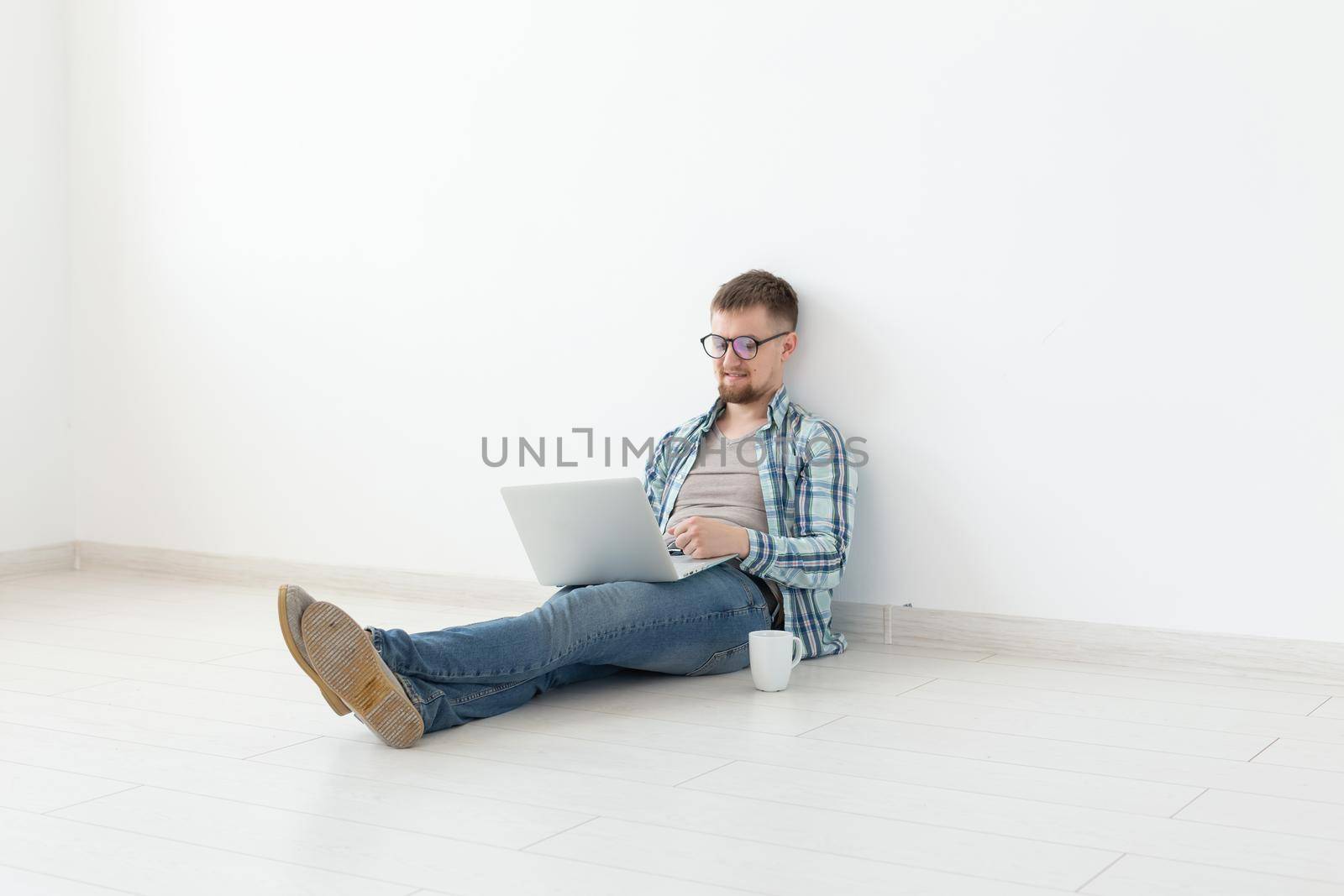 Positive young man in casual clothes surfing the Internet in search of new housing sitting on the floor in an empty room. The concept of finding an apartment using the Internet and a laptop. Copyspace