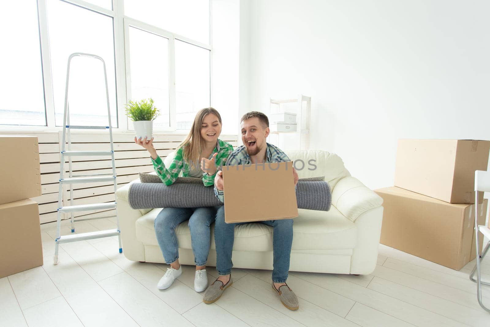 Cheerful joyful young couple charming girl and handsome man holding a box with things and a pot with a plant while moving to a new apartment. Housewarming and mortgage concept. by Satura86