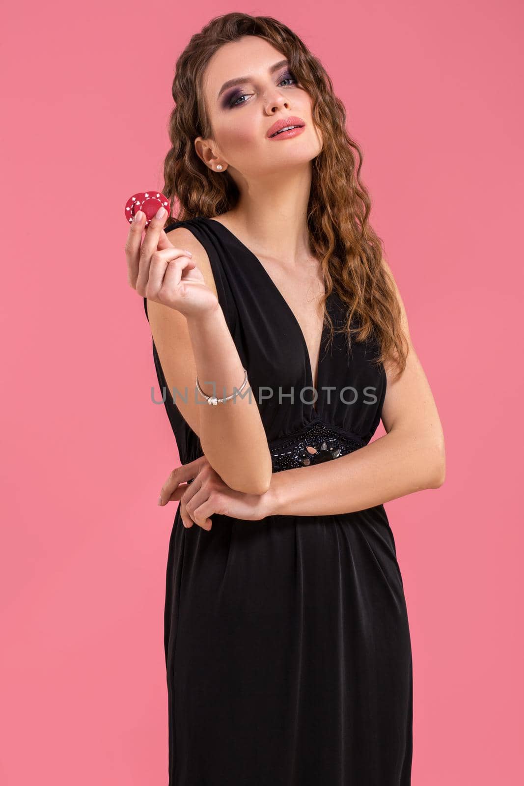 Pretty long hair woman in black dress holding chips for gambling on pink background. Studio shot