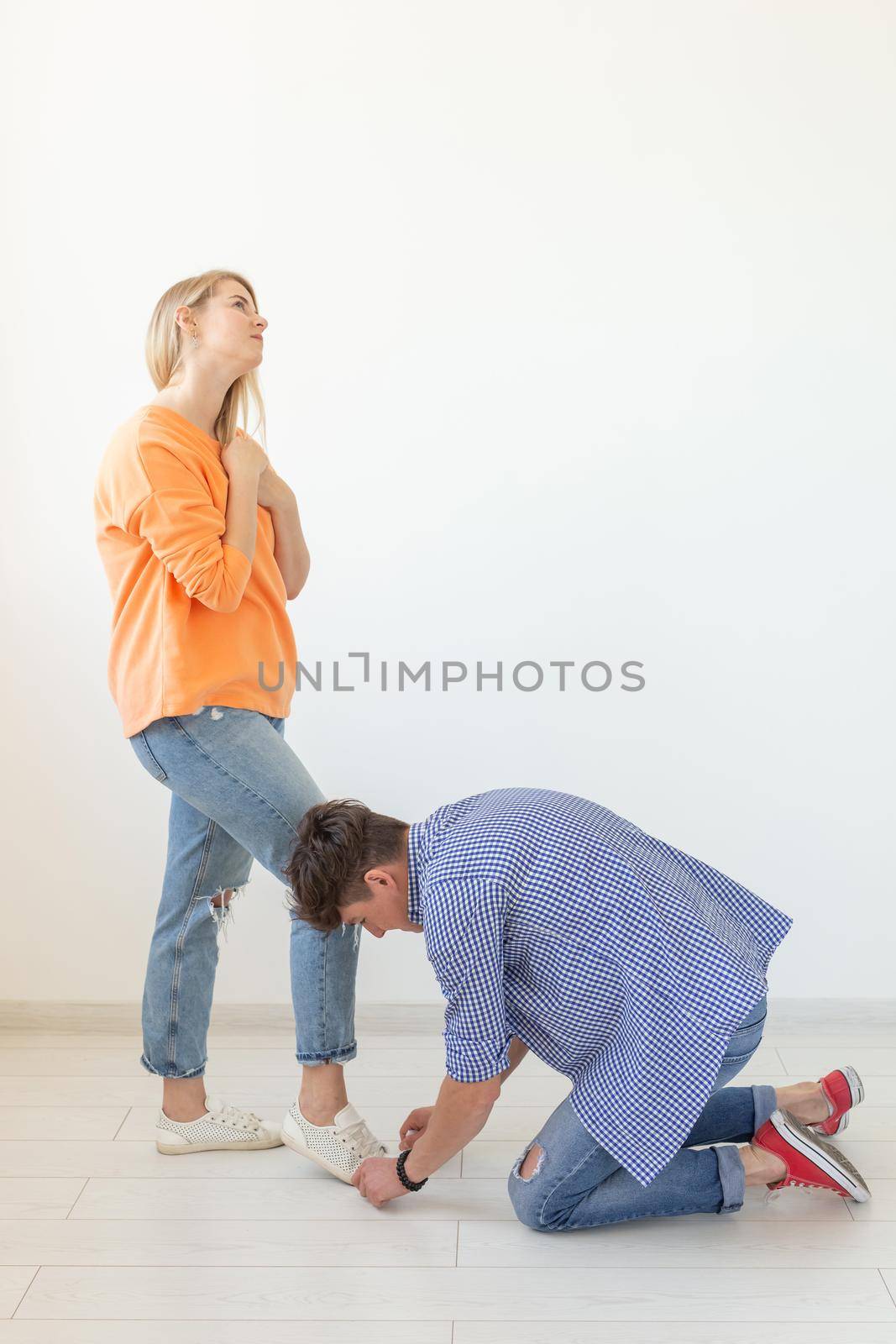 Young man is kneeling and reverently tying shoelaces to his domineering unidentified woman posing on a white background. Concept of dominant relationships. by Satura86