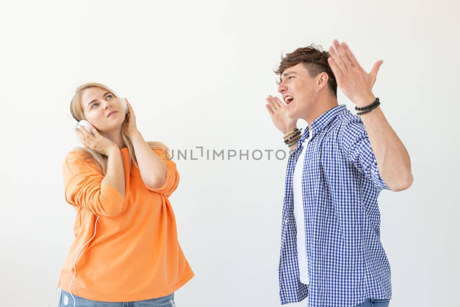 Young upset man begs his woman to listen to him but she listens to music with headphones posing on a white background. Misunderstanding and unwillingness to engage in dialogue. by Satura86