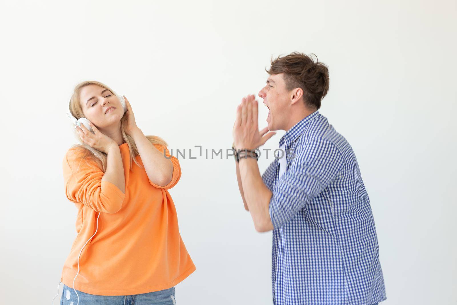 Young upset man begs his woman to listen to him but she listens to music with headphones posing on a white background. Misunderstanding and unwillingness to engage in dialogue. by Satura86