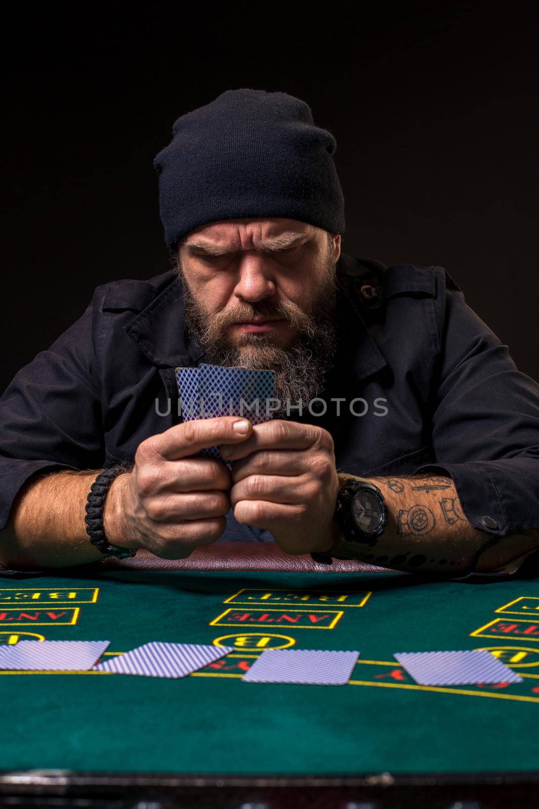 Serious bearded man in a dark blue hat sitting at poker table and holding cards isolated on black. The concept of gambling