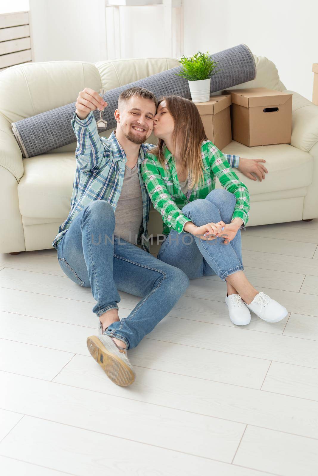 Charming young woman kisses her husband holding in hands the keys to their new apartment while sitting in her new living room. Housewarming and mortgage in new building concept. by Satura86