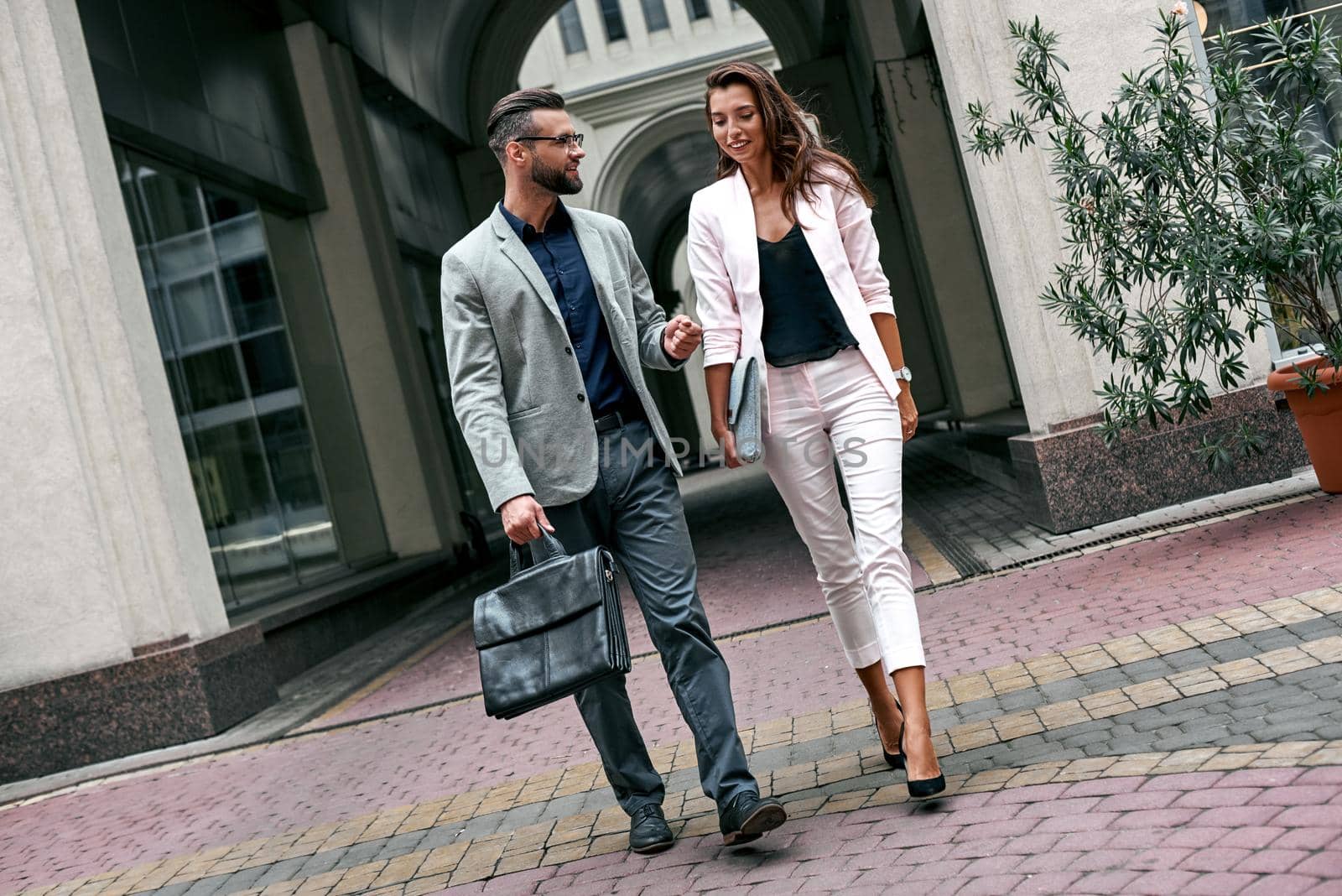 Discussing project. Two young business people walking outside on the city street talking smiling happy man carrying briefcase by friendsstock