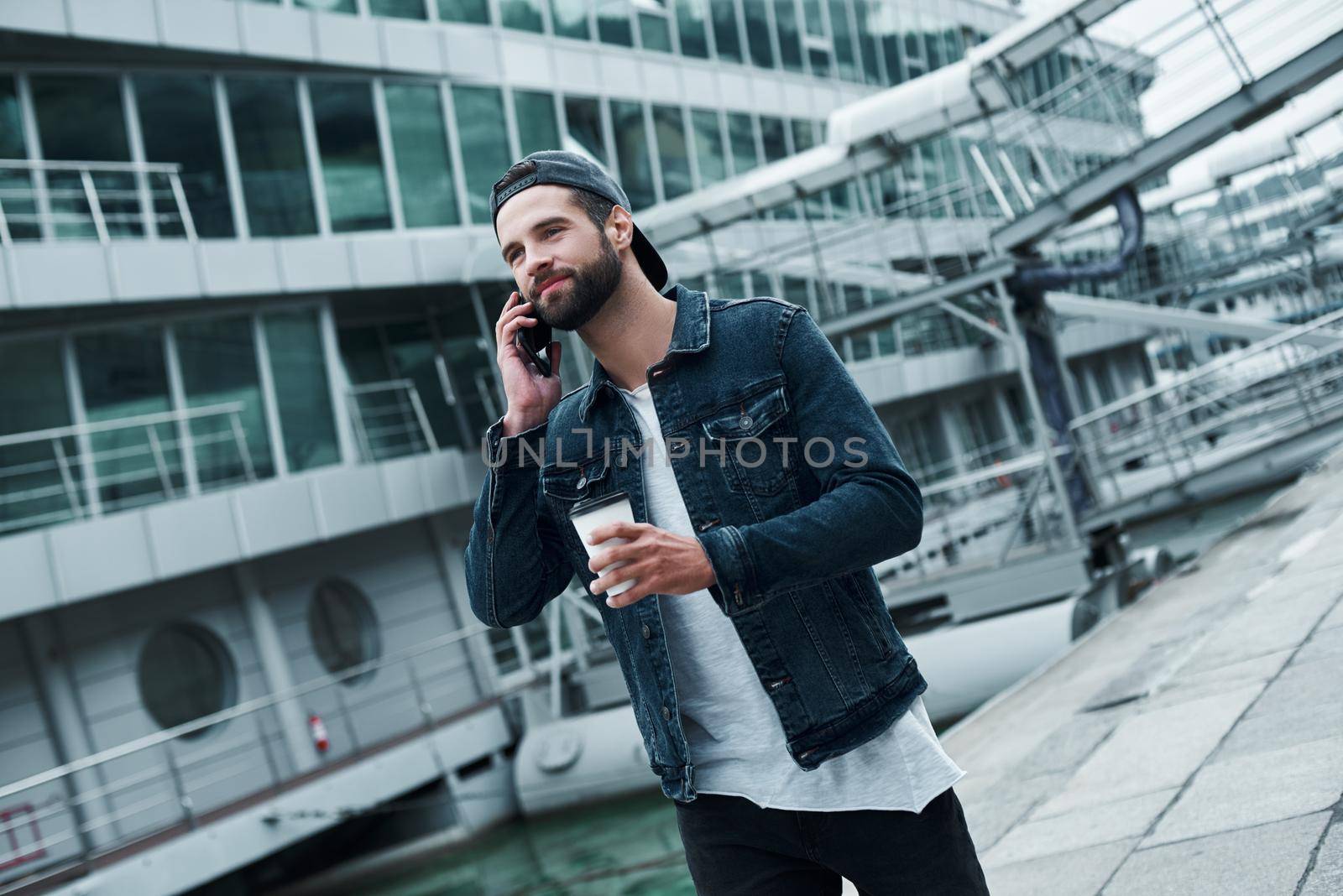 Outdoors leisure. Young stylish man walking on city street talking with friend on smartphone drinking hot coffee smiling joyful