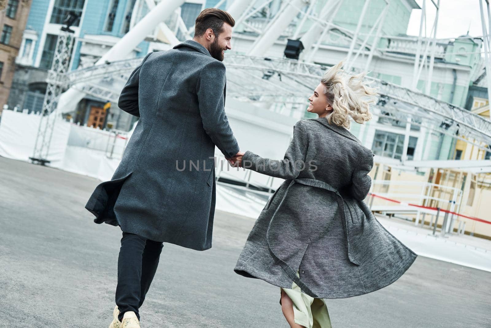 Romantic date outdoors. Young couple running at entertainment park holding hands back view looking at each other smiling happy