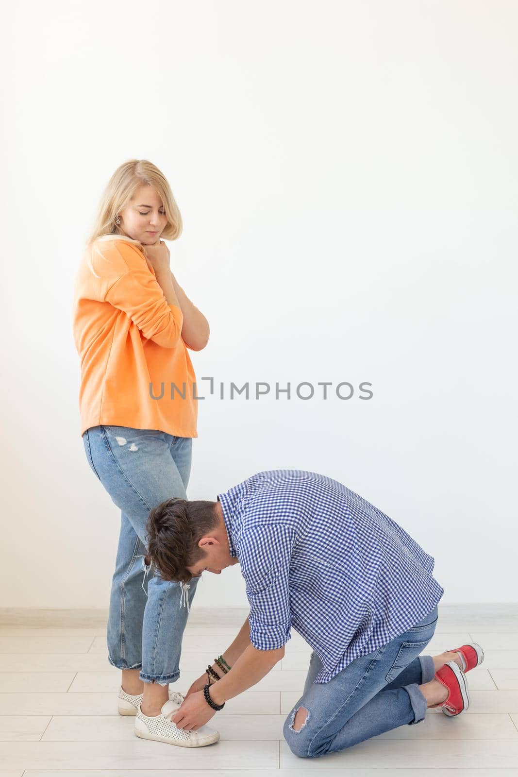 Young man tying shoelaces of his beloved woman posing on the white background. Concept of courtship and reverent relationships. Advertising space