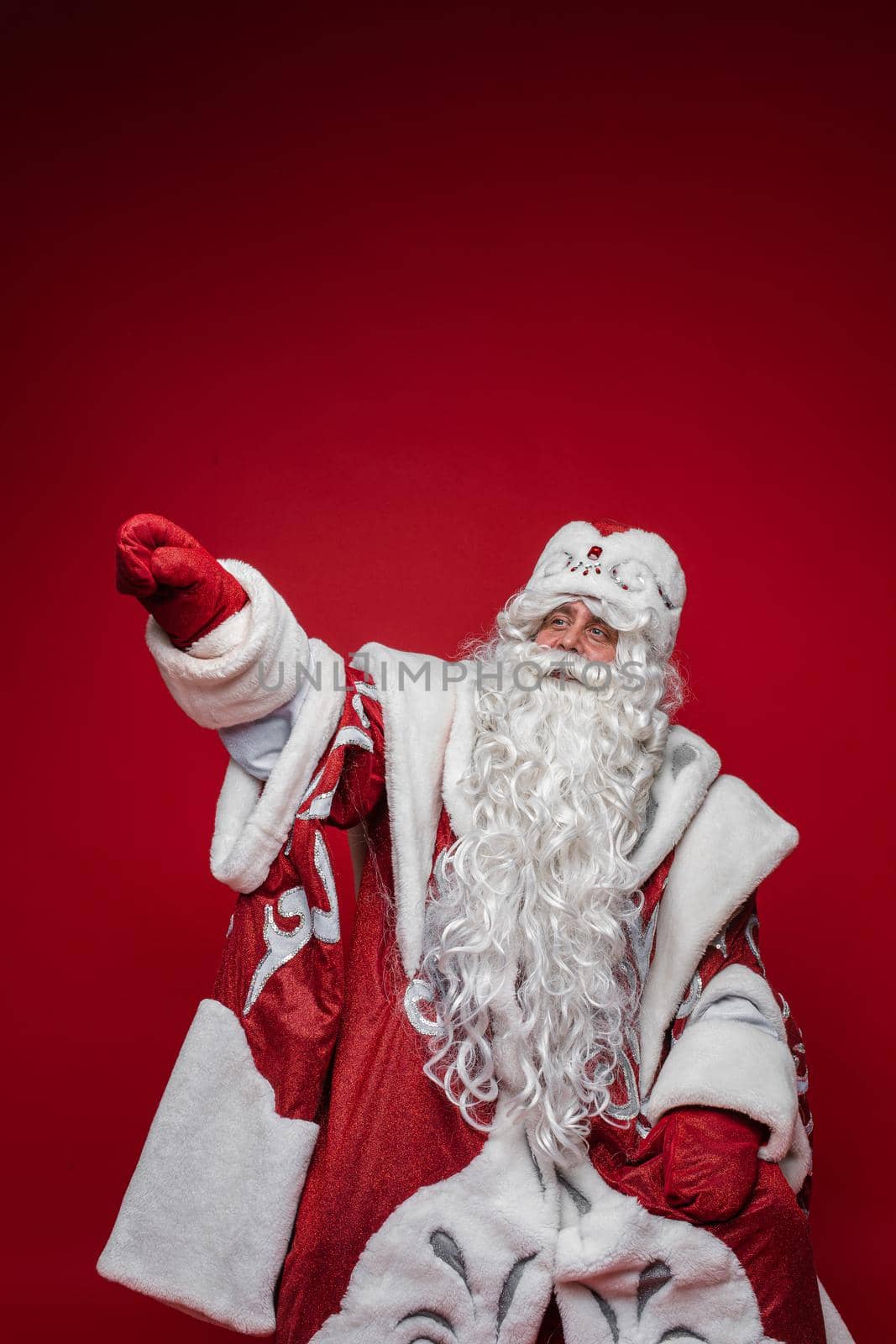 Senior Santa Claus with gray beard pointing by hand away on red background with copy space by StudioLucky