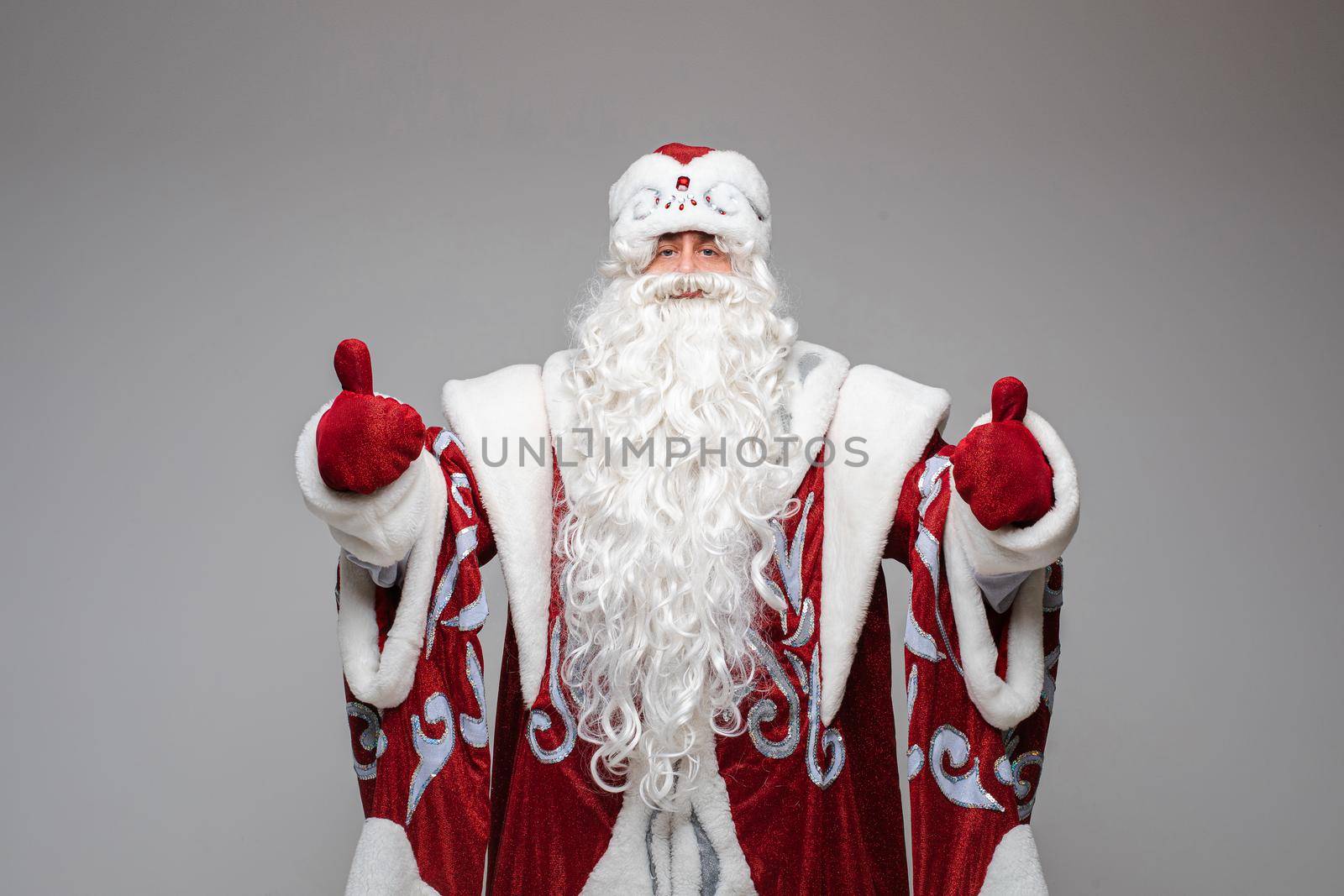 Stock photo of white-bearded Father Frost in red and white hat and traditional costume with red mittens showing both thumbs up.