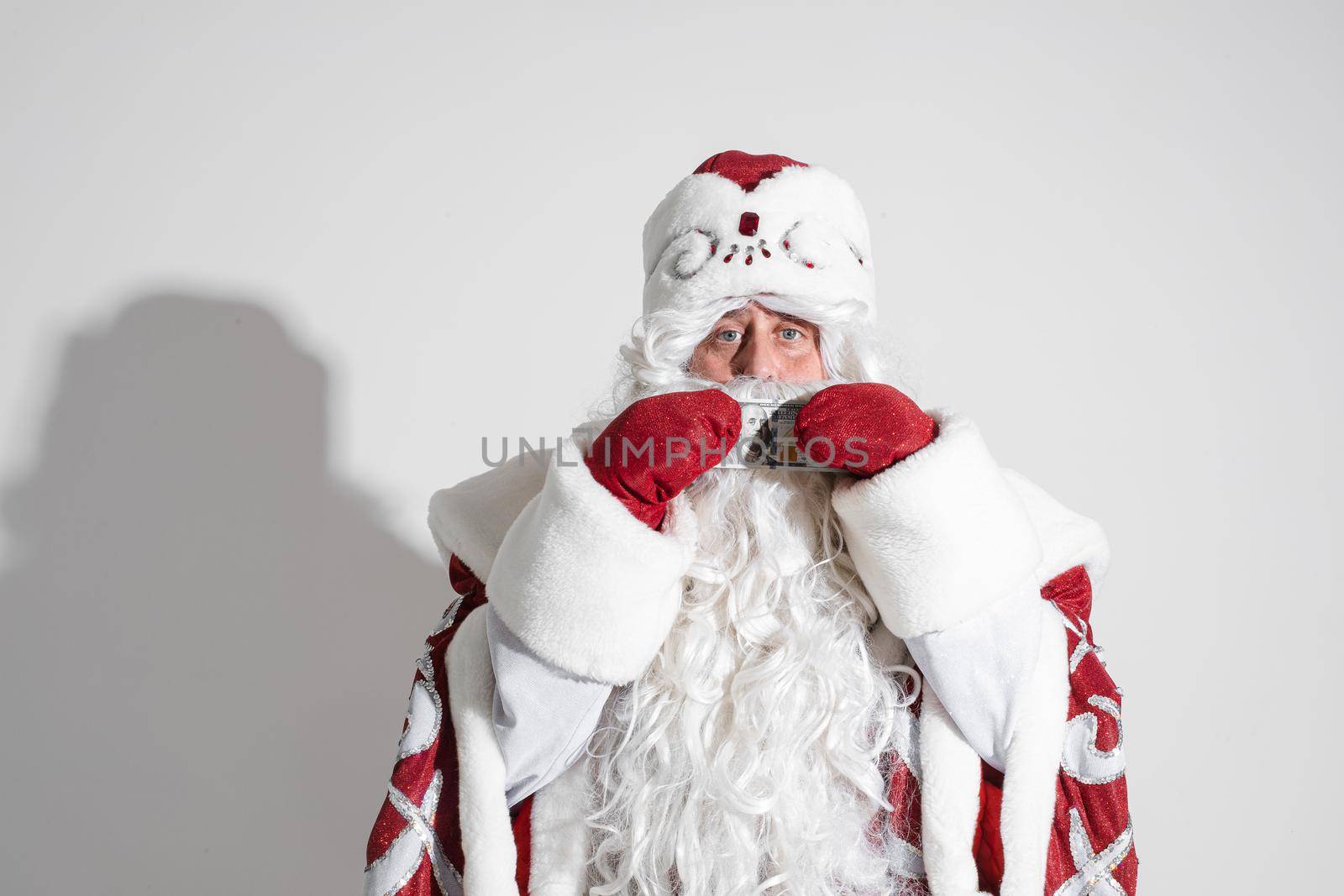 Smiling generous Santa Claus with long white beard in fancy costume holding banknote in hands wearing red mittens. Cutout on white background.