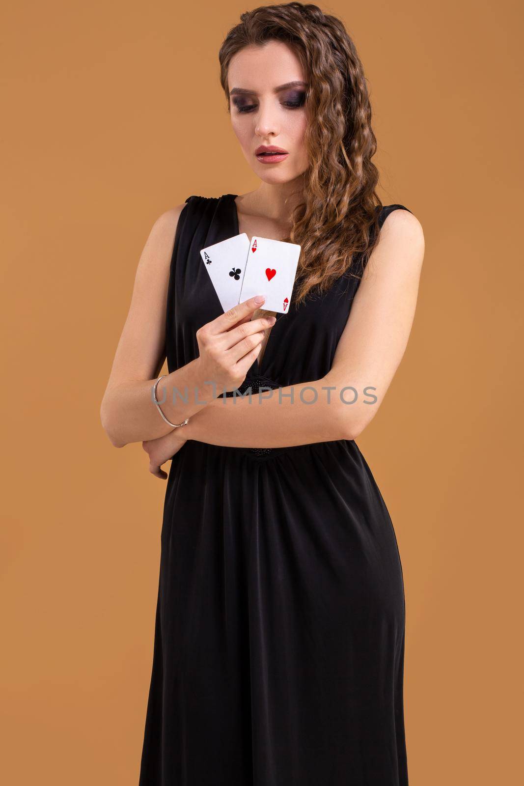 Beautiful brown-haired woman in black dress holding two aces as a sign for poker game, gambling and casino on a beige background. Studio shot