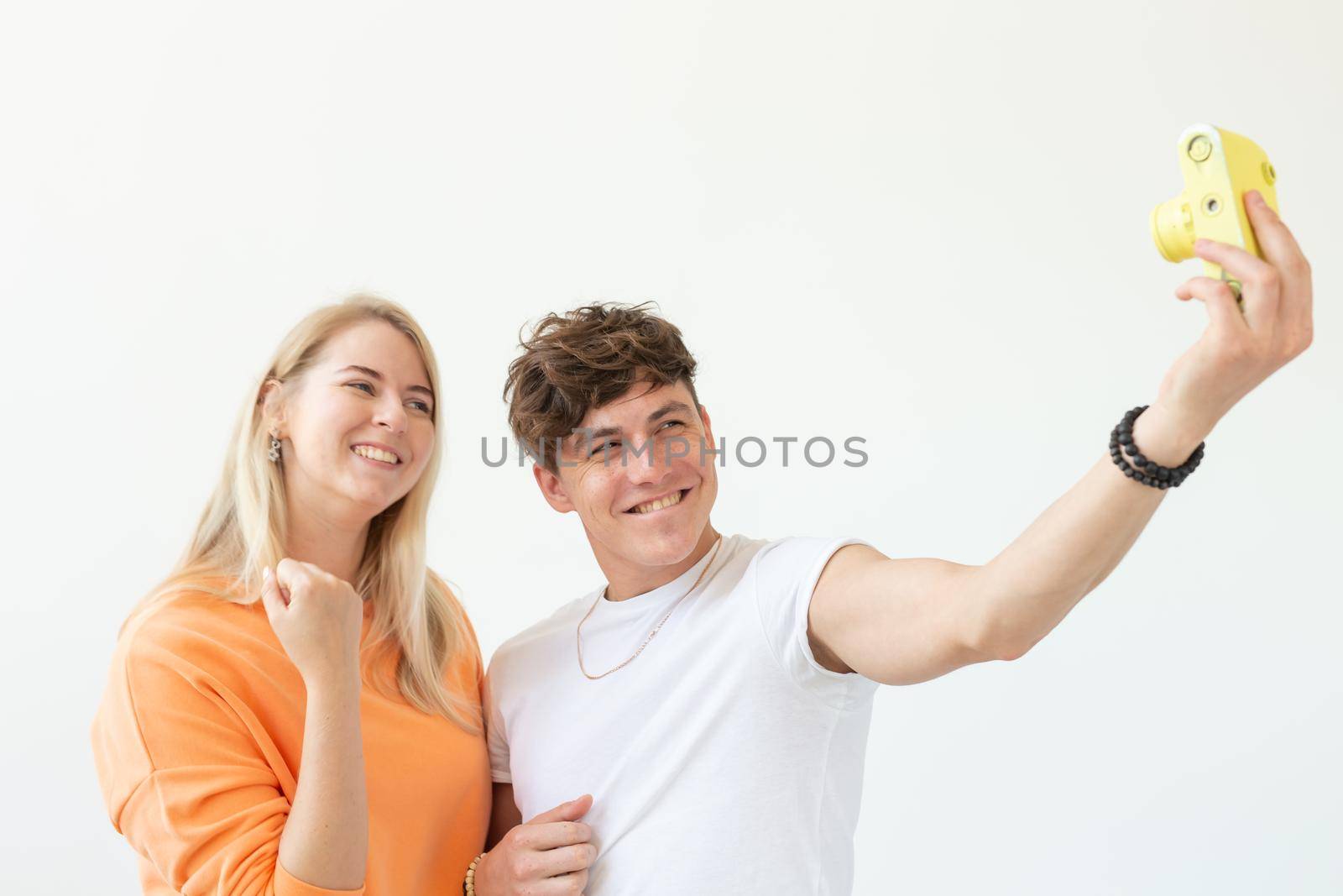 Funny crazy young couple blonde girl and a hipster guy taking a selfie on a vintage yellow film camera posing on a white background. Concept of photography hobby. Advertising space