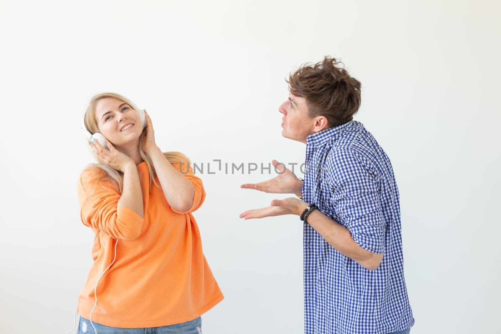 Young upset man begs his woman to listen to him but she listens to music with headphones posing on a white background. Misunderstanding and unwillingness to engage in dialogue