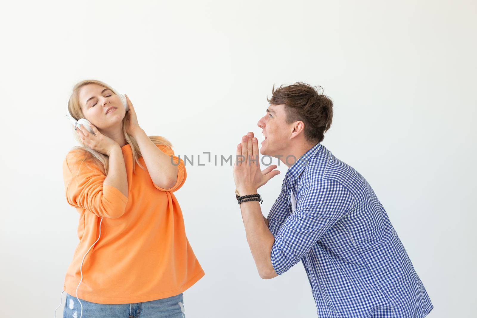Young upset man begs his woman to listen to him but she listens to music with headphones posing on a white background. Misunderstanding and unwillingness to engage in dialogue