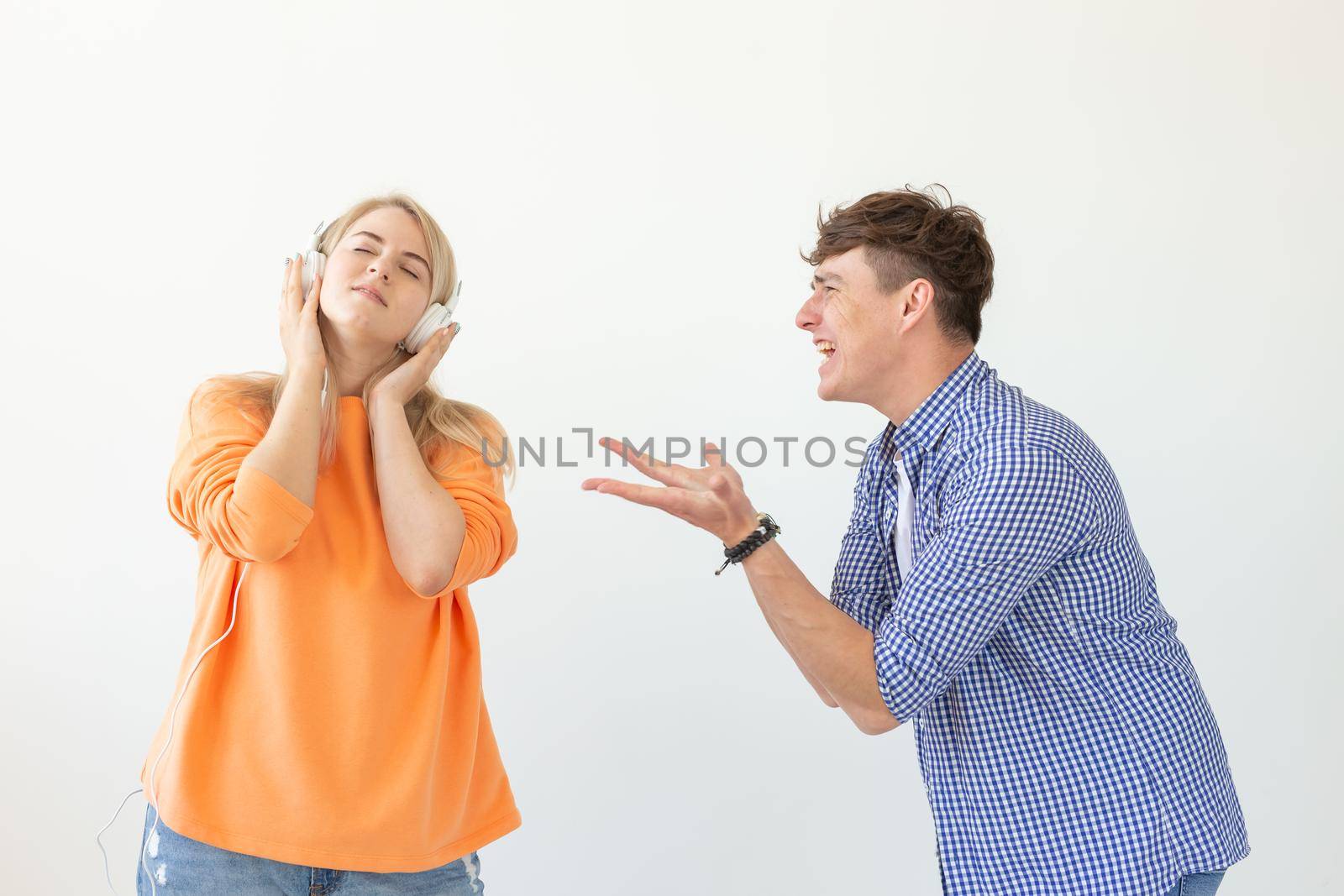 Young upset man begs his woman to listen to him but she listens to music with headphones posing on a white background. Misunderstanding and unwillingness to engage in dialogue