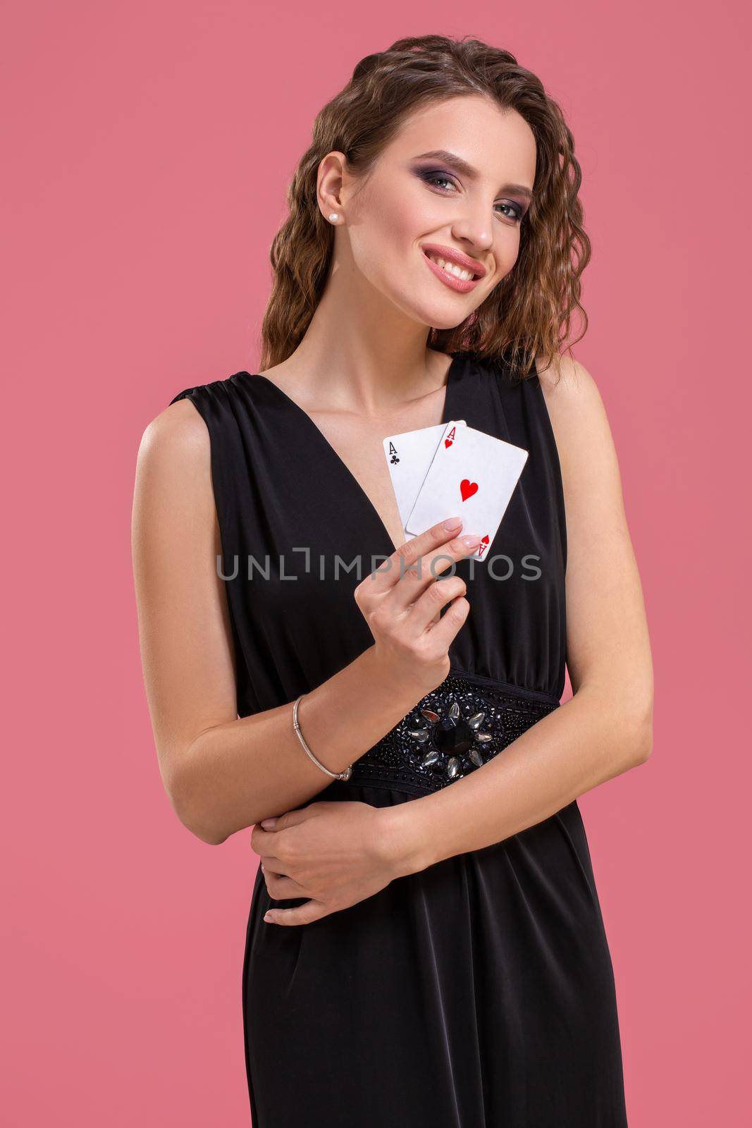 Beautiful caucasian woman in black dress with poker cards gambling in casino. Studio shot on a pink background