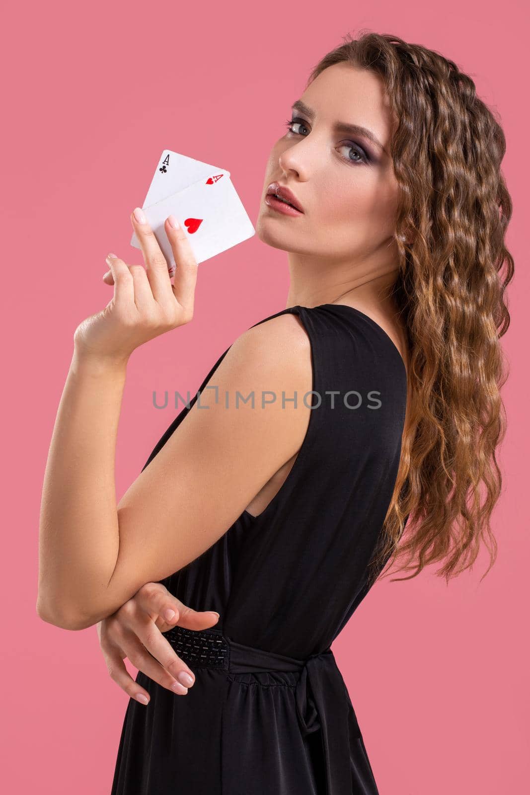 Young woman in black dress holding two aces in hand against on pink background. Studio shot