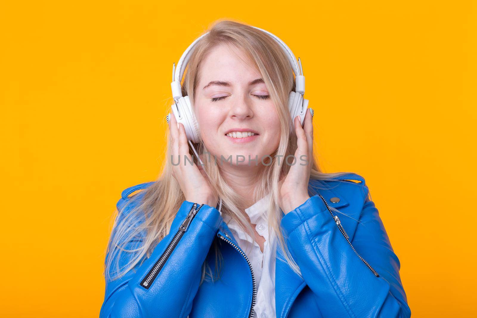 Portrait of pretty young girl blonde female student holding smartphone with blue leather jacket headphones posing on a yellow background. Concept of listening to online radio and music subscription