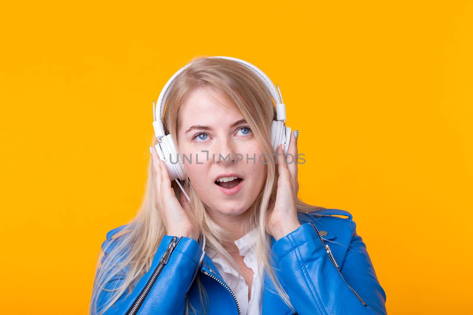 Portrait of pretty young girl blonde female student holding smartphone with blue leather jacket headphones posing on a yellow background. Concept of listening to online radio and music subscription